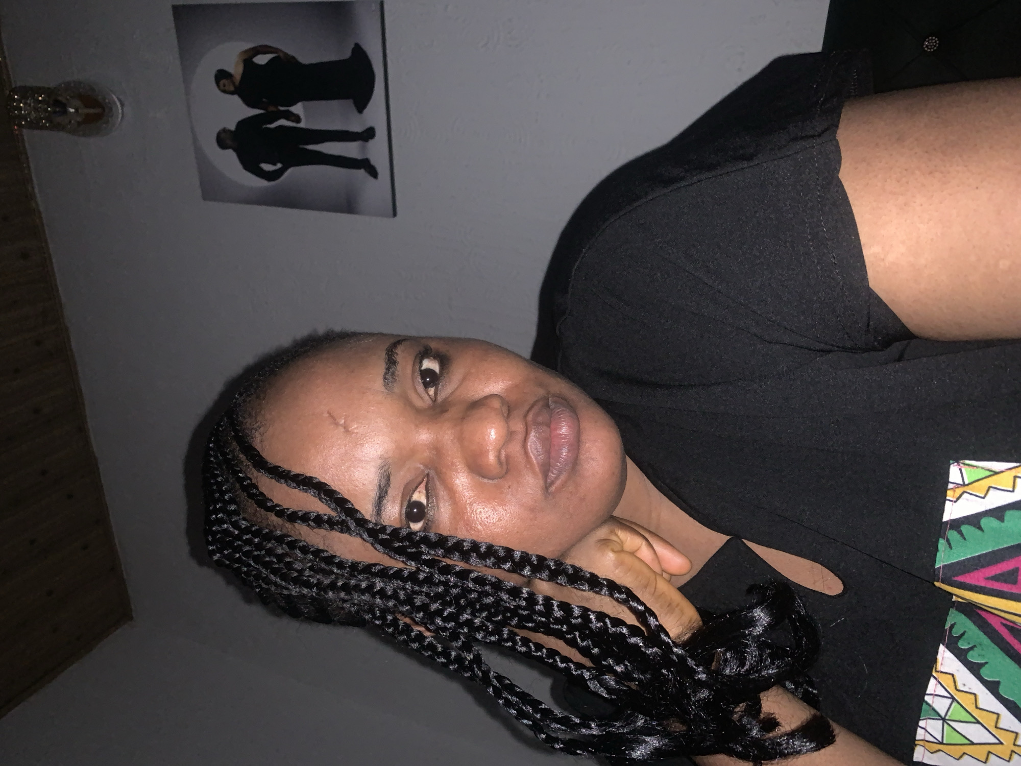 A medium close-up shot shows a Black woman from the chest up, with her head tilted to her right, resting her chin on her left hand. Her hair is braided and hangs over her left shoulder. She is wearing a black t-shirt with a colorful, patterned trim around a V-shaped neckline. The background is a gray wall with a framed picture of a couple in formal attire hanging on it, and a decorative item resembling a high-heeled shoe on the ceiling. The lighting is dim, with the woman's face illuminated from the front. The image appears to have been taken indoors in a domestic setting.