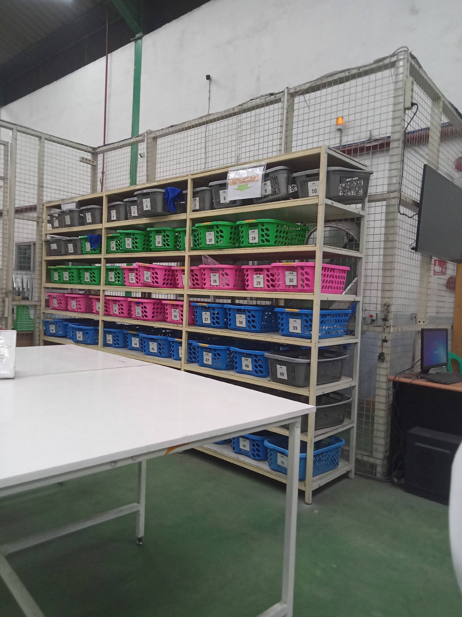 The image depicts a storage area within what appears to be a manufacturing or distribution facility. The primary focus is on a multi-tiered shelving unit filled with an assortment of plastic bins and baskets. The shelves are organized with bins of various colors, including gray, green, pink, and blue, many of which are labeled with numbers and text such as "LINE" followed by a number. On the top shelves, several gray bins with lids are visible. Below these are rows of green bins, followed by pink bins, and then blue bins. The shelves themselves are made of a light-colored metal frame.
To the left of the shelving unit, a white table with metal legs is partially visible. The floor is a light green, consistent with industrial flooring. In the background, a wire mesh cage structure encloses part of the facility. A small orange warning light is mounted on top of this structure. Further to the right, a black computer monitor sits on a desk, accompanied by a computer tower and a green chair. The walls are painted white, and there are visible conduits and electrical wiring.
The image is taken from a low-angle perspective, looking slightly upwards towards the shelving unit. The lighting appears to be artificial, common in indoor industrial settings. There are no people visible in the scene. The overall impression is one of an organized and functional storage system within a production environment.