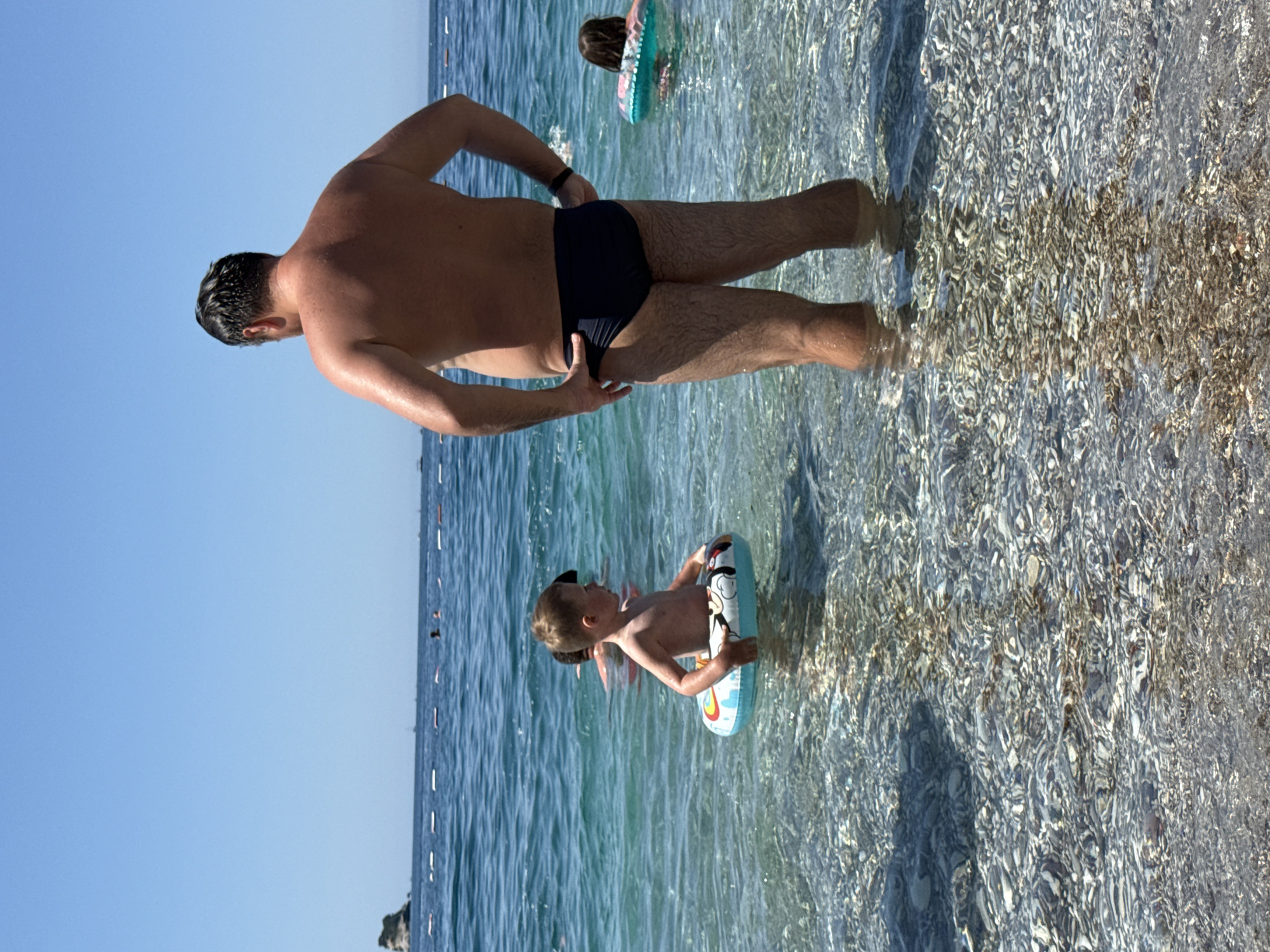 This image depicts a sunny beach scene in Budva, Montenegro. In the foreground, an adult male stands in clear, shallow water with his back to the viewer, wearing dark-colored swim briefs and with his hands resting on his hips. His hair is dark and appears wet. To his left, a young boy, appearing to be a toddler or preschooler, stands partially submerged in the water while wearing a blue and white inflatable swim ring with a Mickey Mouse design. The boy is shirtless and faces towards the right side of the frame. Further out to the right of the adult, another child is visible from behind, also in the water with a light blue inflatable swim ring. The water is highly transparent, revealing a seabed composed of numerous small, light-colored pebbles and stones. A line of white and red buoys extends horizontally across the middle ground, indicating a swimming area boundary. Beyond this line, several dark silhouettes of individuals are visible in the water, and multiple small boats or yachts are present on the distant horizon. A distinct rocky islet is visible on the far left side of the horizon. The sky is clear and bright blue.