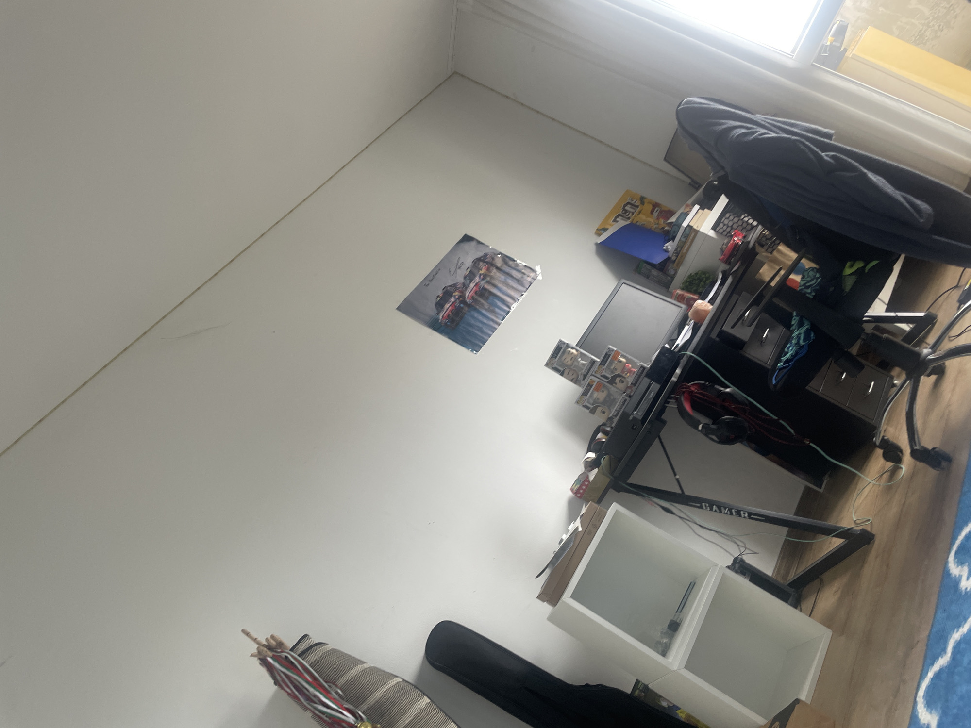 This image depicts an interior room in Sofia, Bulgaria, viewed from an elevated, angled perspective towards a corner. The walls are white, and the floor features light-colored wooden planks, partially covered by a blue rug with white patterns in the lower right foreground. Natural light enters from a window positioned high on the right wall.
A black gaming desk is situated against the right wall. On its surface, a black computer monitor is visible, along with several stacked Funko Pop figures. A pair of black headphones with red ear cup accents and associated red and teal cables are draped over equipment on the desk. A black office chair, covered with a dark blue or black garment, is positioned in front of the desk. Above the desk, a blue folder and various papers or posters are visible. A prominent "GAMER" branding is affixed to a support bar of the desk. Beneath the desk, a white modular shelving unit with two open cubbies contains a few small items.
On the left wall, a poster depicting multiple racing cars is affixed. In the lower-left foreground, a striped cushion is visible, supporting a red, white, and green striped object, consistent with a folded umbrella. Adjacent to this, a black soft case, possibly for an instrument, rests on the wooden floor. The room appears unoccupied.