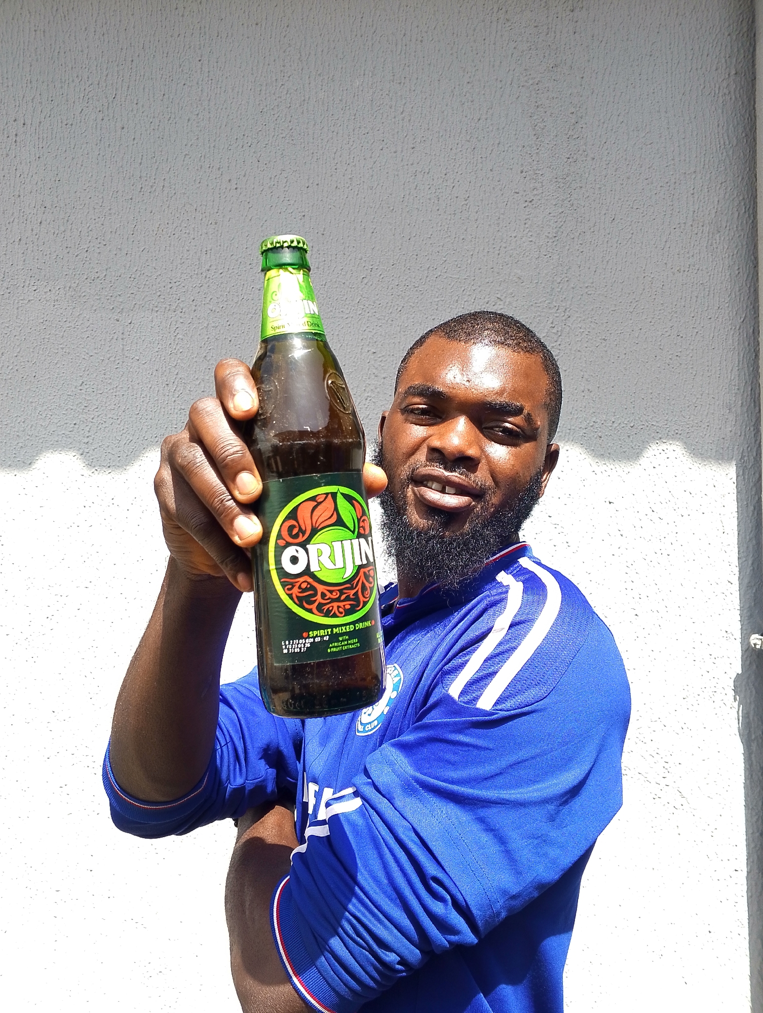 A man with dark skin and a beard holds up a bottle of Origin Spirit Mixed Drink towards the camera. He is wearing a blue athletic shirt with white stripes on the sleeves. The background is a textured white wall, with shadows cast by sunlight. The man is looking at the camera with a slight smile. The bottle's label is visible, featuring the brand name "ORIJIN" in large green letters within a circular emblem. Text on the label indicates it is a "SPIRIT MIXED DRINK" with "AFRICAN HERBS & FRUIT EXTRACTS." The image appears to be taken outdoors during the daytime, likely in a sunny environment, suggested by the bright lighting and the shadows. The overall mood is casual and perhaps celebratory.