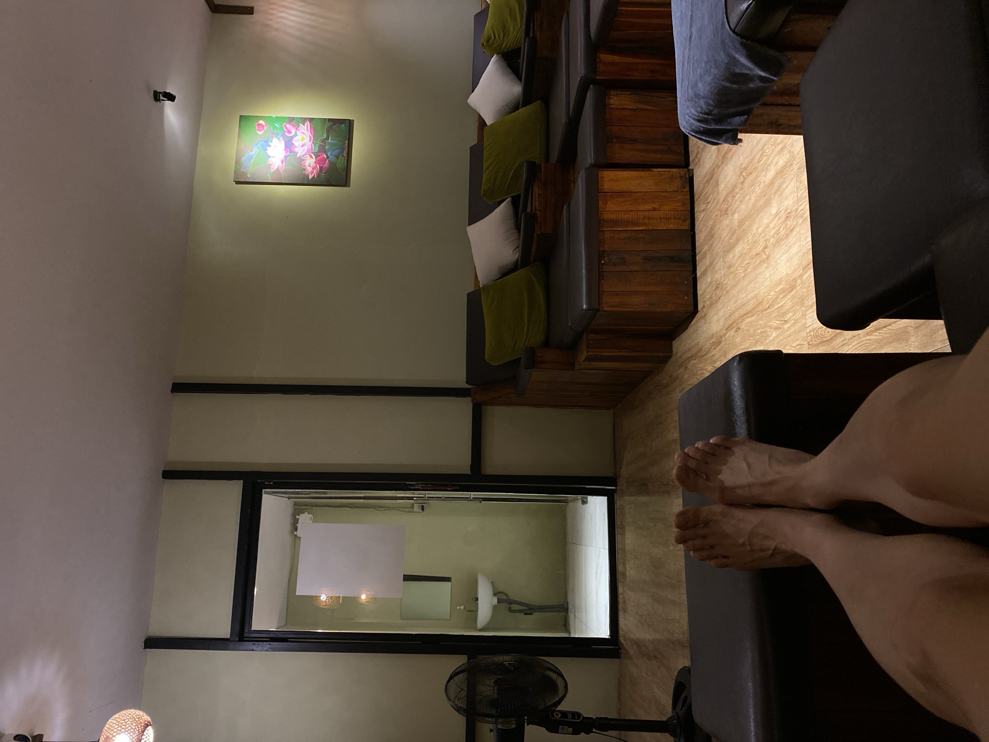 The image depicts the interior of what appears to be a spa or massage parlor, possibly in Nha Trang Ward, Vietnam. The scene is oriented sideways. In the foreground, a person's legs and bare feet are resting on a dark, cushioned surface, suggesting they are a client receiving a service. The room features tiered seating upholstered in dark material, with wooden accents on the sides, arranged in a lounge-like setting. Several green and white cushions are placed on the seating.
To the left, on a cream-colored wall, a framed artwork of pink lotus flowers is illuminated by a spotlight mounted above it. Further to the left, a lampshade with a woven texture is visible, casting a warm glow.
In the background, a large glass-enclosed area reveals a small washroom or changing area. Inside, a white sink is mounted on the wall, with plumbing visible. A white board or sign is also affixed to the wall within this area. A black oscillating fan stands in front of this glass enclosure. The flooring in the main seating area is a light-colored wood-grain laminate, while the floor in the enclosed washroom appears to be tiled. The overall ambiance suggests a relaxing and tranquil environment. There is no visible text in the image. The time of day is indeterminate, but the lighting suggests it could be evening or indoors with artificial lighting.