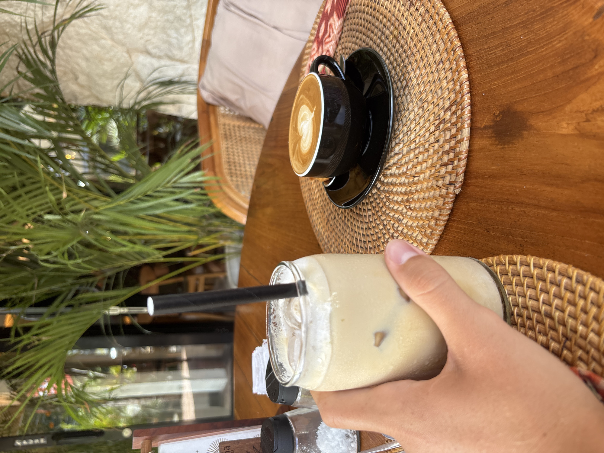 A first-person, high-angle shot shows a hand holding a glass of iced coffee with a black straw. A black cup of coffee with latte art sits on a woven placemat on a wooden table. Outside, green tropical foliage is visible. The scene is in Kuta Selatan, Indonesia.