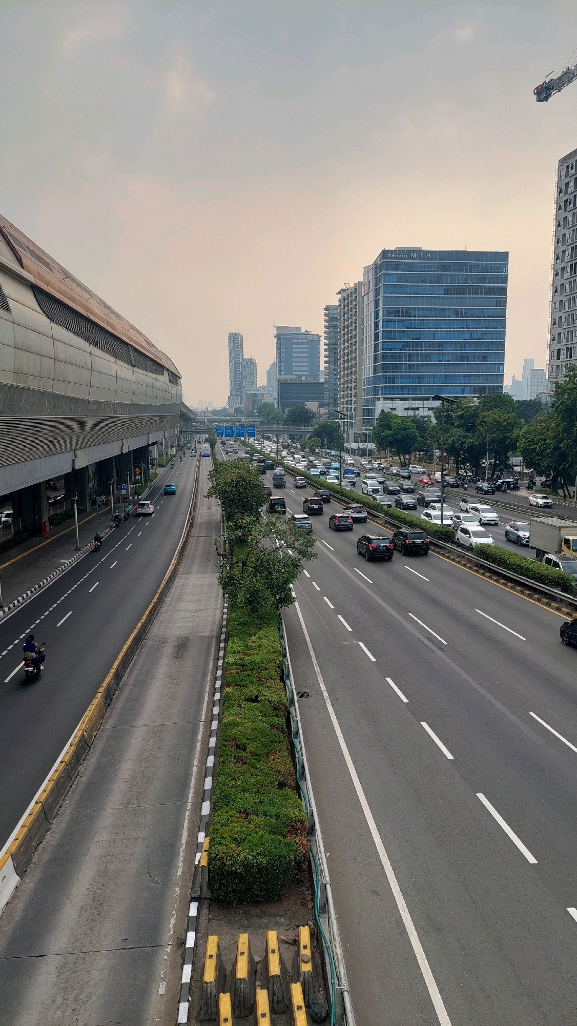 The image shows a multi-lane highway in South Jakarta, Indonesia, with significant traffic congestion. A modern elevated train station runs along the left side of the highway, featuring a sleek, metallic facade and a reddish-brown roof. The highway itself is divided by a green median strip with manicured bushes and trees. Numerous cars, motorcycles, and a few trucks are visible on the road, moving in both directions. Several tall modern buildings, characteristic of a city skyline, dominate the background, including a prominent blue glass skyscraper labeled "Menara MTH". The sky is overcast, suggesting a cloudy or hazy day. On the pedestrian walkway or overpass overlooking the highway, there are concrete barriers and some yellow traffic cones.