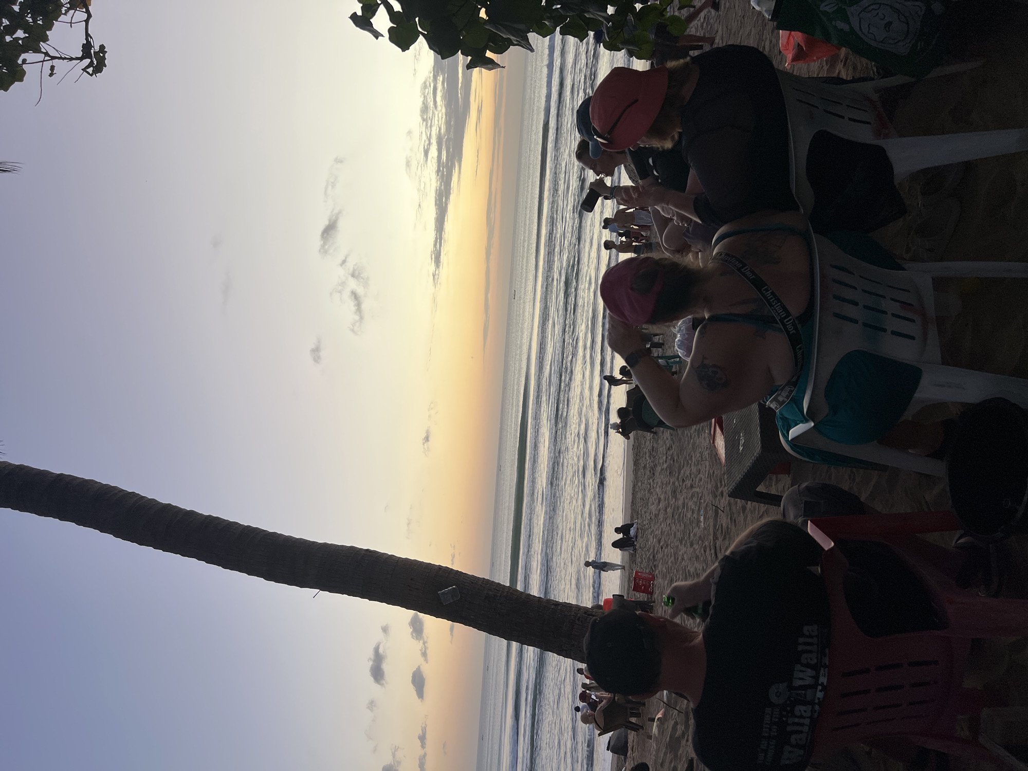 This image captures a sunset scene on Kuta Beach, Bali, Indonesia. The sky is painted with soft hues of orange, yellow, and purple, with scattered clouds. The ocean waves roll gently onto the sandy shore, where several people are gathered. In the foreground, four individuals are seated on chairs, facing the ocean. One person in the front left is wearing a black t-shirt and a black baseball cap, holding a green bottle. To their right, a person wearing a pink hat and a blue top is visible from the back. Next to them, another person in a red hat and black top is seated. Further to the right, a fourth person is partially visible, also seated.
Further back on the beach, other beachgoers are seen strolling, sitting, and interacting. A tall palm tree stands prominently in the left half of the frame, leaning slightly. In the distance, the horizon is dotted with a few small boats. The scene is characteristic of a relaxed evening on a popular tourist beach.