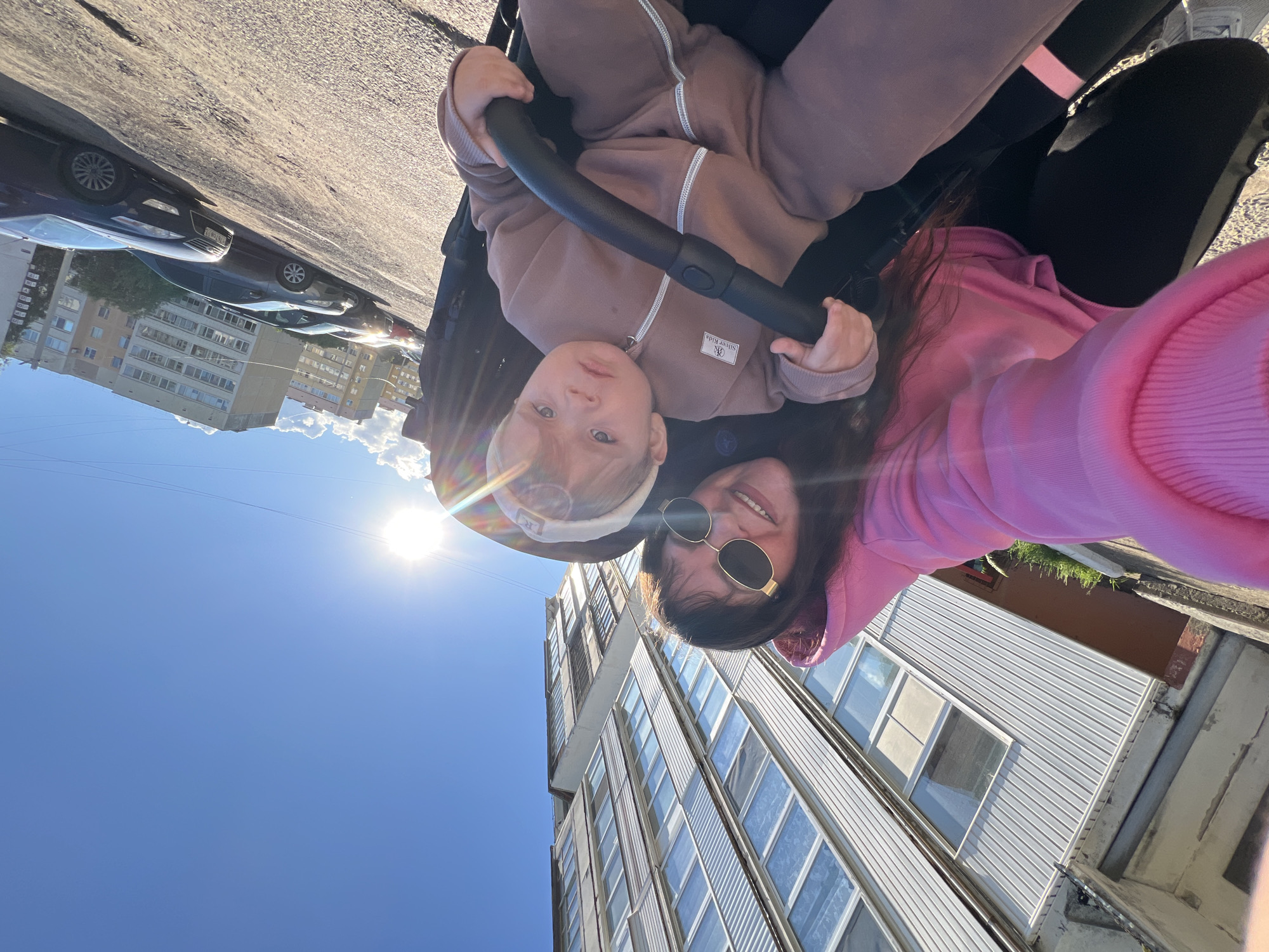 A selfie shows a young woman and a baby in a stroller, taken outside in Noviy Urengoy, Russia. The woman is wearing a pink hoodie and sunglasses, while the baby is in a brown onesie and gripping the stroller's bar. The sun is visible in the sky, and a tall, light-colored apartment building is in the background. Other buildings and parked cars are also visible.