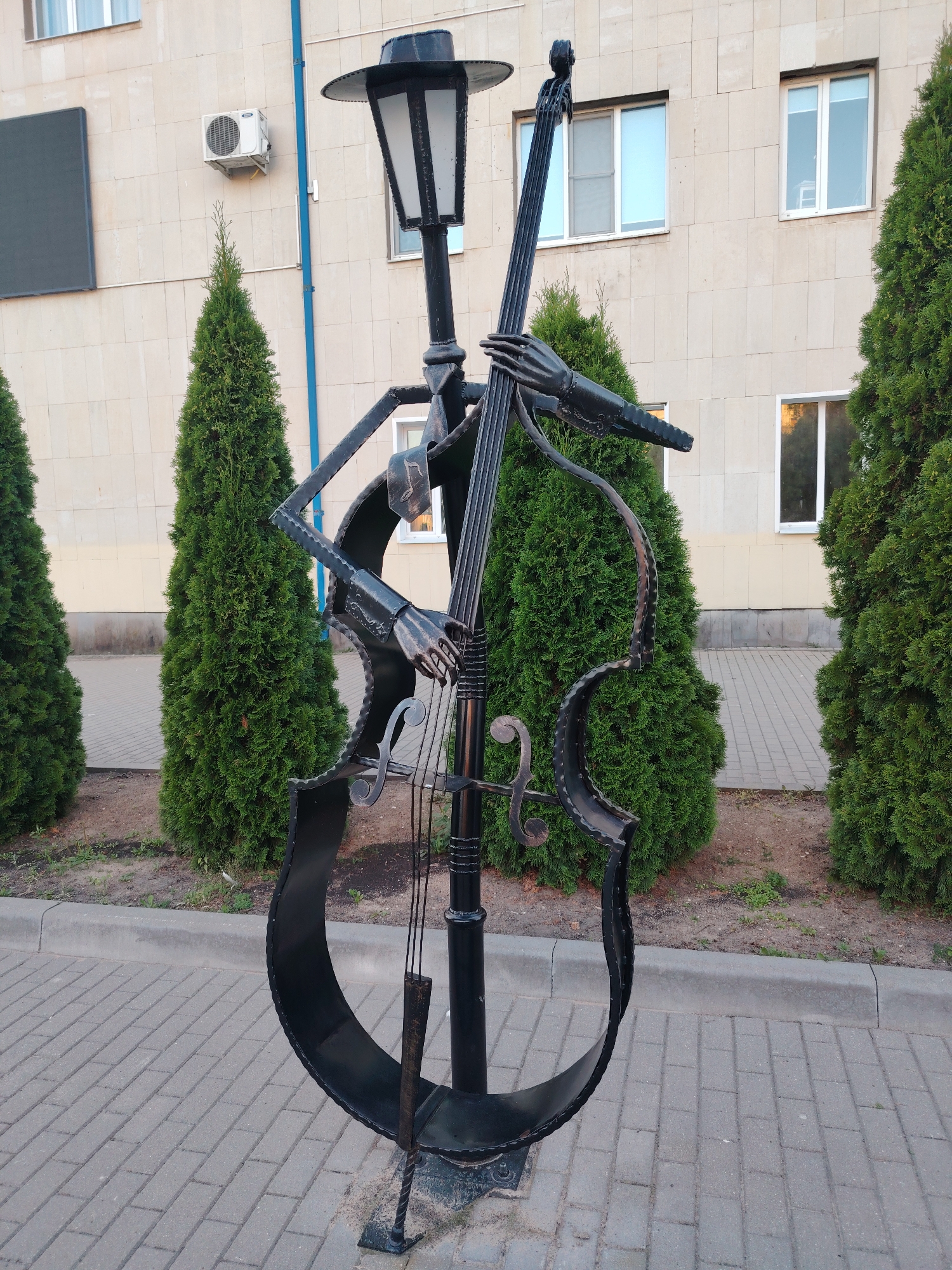 A black metal sculpture resembling a cello and a lamppost is situated on a paved walkway in front of a building. The sculpture depicts a stylized figure playing the cello, with the lamppost integrated into the instrument's neck. Several evergreen trees flank the sculpture. The building behind it has a light-colored, stone-like facade with multiple windows and an air conditioning unit mounted on the exterior wall. The scene appears to be in a public outdoor space.
