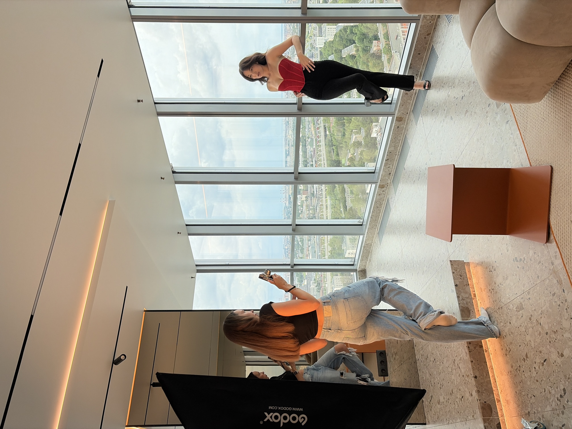 Two young women are posing for a photoshoot in a modern apartment with large windows overlooking a city skyline. In the foreground, a woman with long brown hair is taking a photo with a smartphone of another woman. She is wearing ripped light-wash jeans and a black crop top. The second woman is standing near the window, leaning on it and looking away from the camera. She is wearing a red corset top and black pants, with her hair down. The apartment features a large mirror reflecting the scene, a black photography reflector, and a small orange side table. The floor is a polished terrazzo material. The backdrop is a panoramic view of a city, with buildings and trees visible under a cloudy sky. Based on the architecture visible, it is plausible this apartment is located in the city of Saint Petersburg, Russia.
