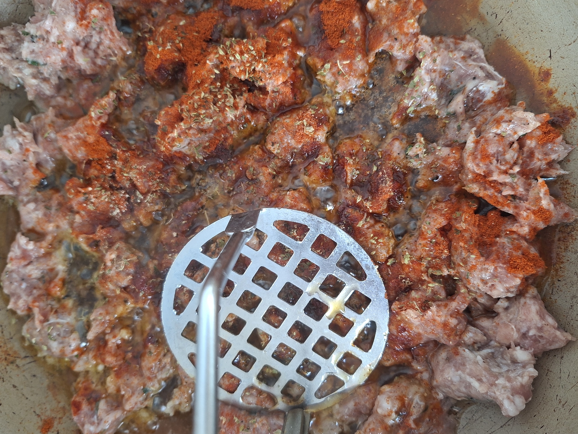 A close-up overhead shot shows ground meat cooking in a pan. The meat is seasoned with a red spice, possibly paprika, and dried herbs. A metal slotted utensil is pressing down on the meat. The pan appears to be non-stick, and there is liquid in the pan. The image does not provide enough information to ascertain its location in Galabovo, Bulgaria.