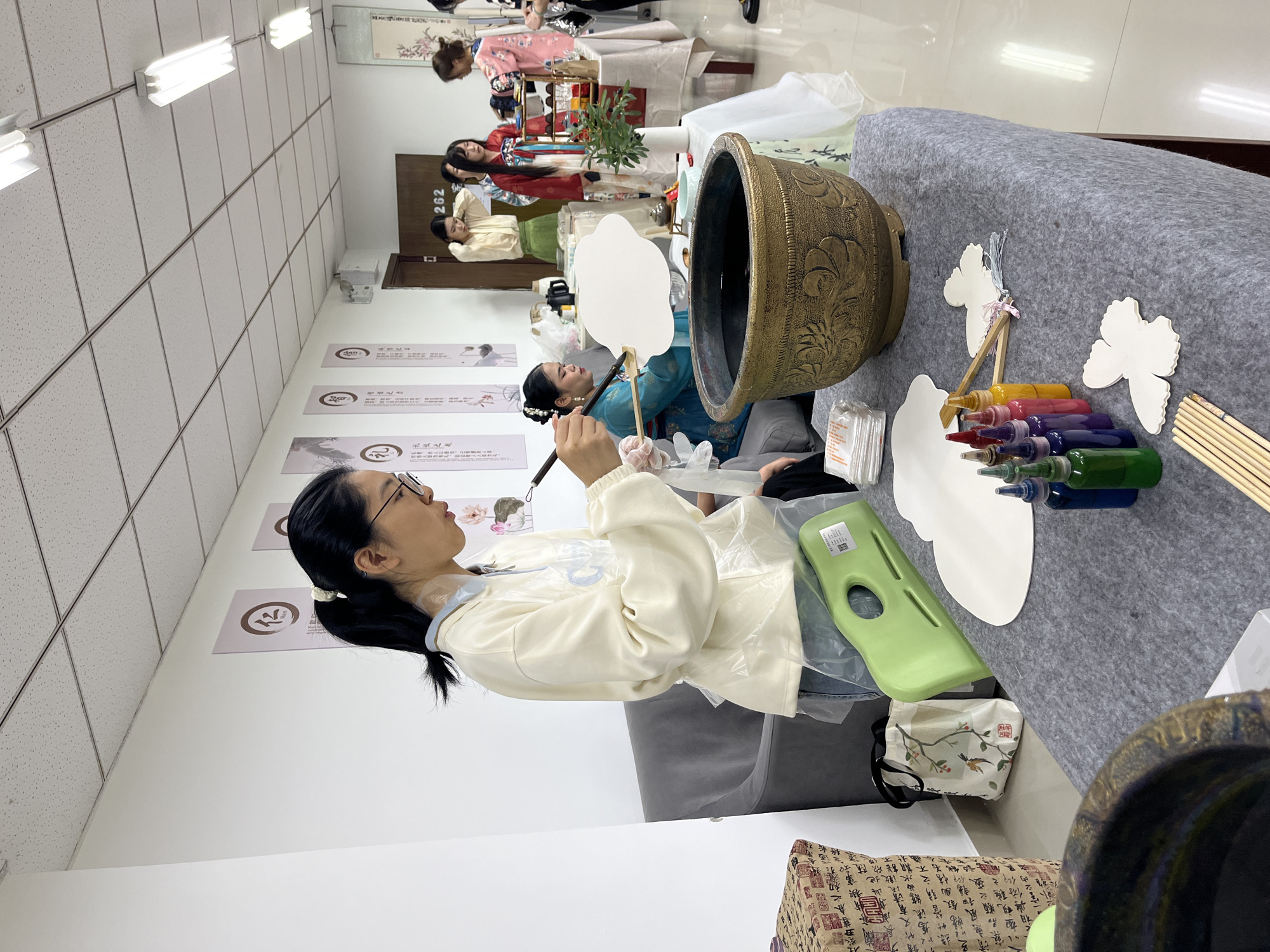 Here is a detailed description of the image:
The image captures an indoor setting, likely a classroom or studio, where a group of women are engaged in an artistic activity. The main focus is on a woman on the left who is wearing glasses and a light-colored jacket. She holds a paintbrush and appears to be painting on a white, fan-shaped object.
Several other women are seated or standing in the background, some wearing traditional Hanfu clothing. They seem to be involved in similar creative endeavors. The room is lined with posters or banners featuring Chinese calligraphy and artwork, suggesting a cultural or educational theme.
In the foreground, a large, ornate pot sits on a table covered with a gray cloth. Nearby are several bottles of colorful liquids, possibly paints or dyes, as well as brushes and more white fan-shaped objects. These items suggest that the women are involved in a painting or dyeing activity.
The location in the city of Jiangjin, China can be inferred from the details present in the image. Without a specific location being provided, I cannot infer the location of Jiangjin, China.