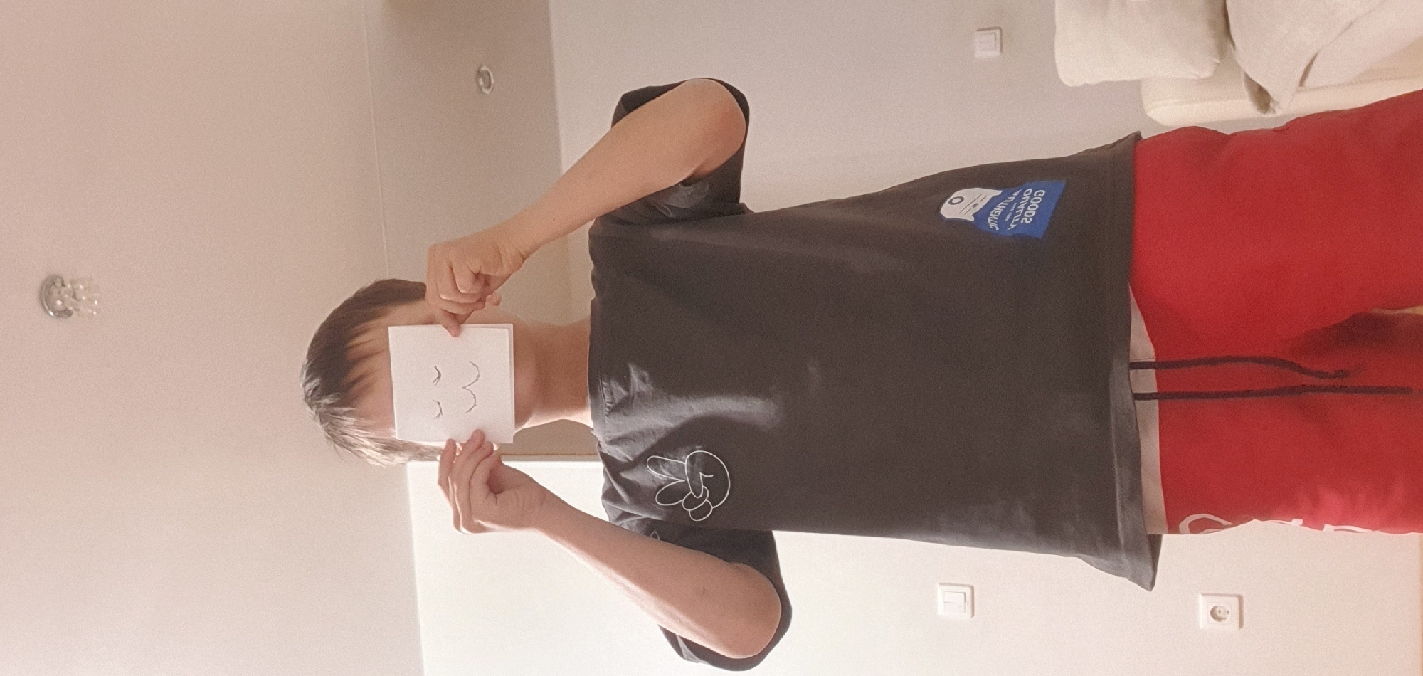 A person stands indoors holding a square piece of paper with curved lines resembling eyelashes drawn on it, covering their face. They wear a dark gray t-shirt with a blue patch and a white peace sign emblem and red sweatpants. The location appears to be a room with a white wall and electrical outlets. Based on the provided context of "Chaikovsky, Russia," it can be inferred that the location is within the city of Chaikovsky, Russia. Furniture and a light fixture are also visible.