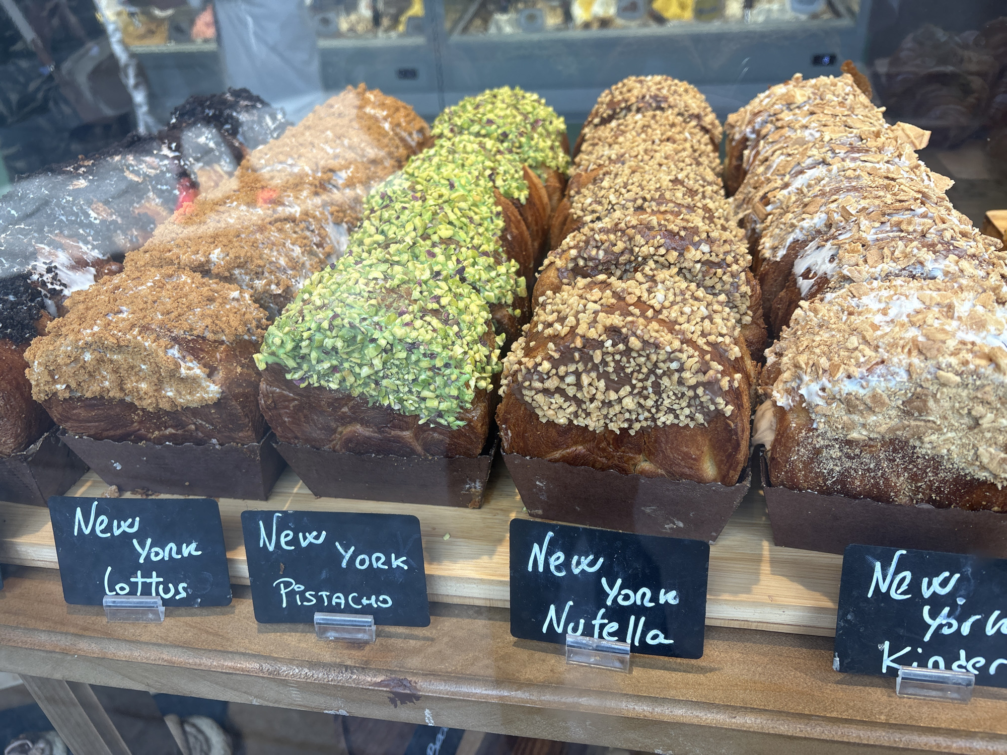 The image shows a display of gourmet pastries in what appears to be a bakery or cafe window. Several large, elaborate pastries are arranged in rows, each one seemingly a different flavor or topping. The pastries are long and rectangular, with a layered, flaky texture. Some are covered in chopped nuts, others in green pistachios, and one has a dark topping that could be chocolate or a dark cookie crumble. There are also pastries with lighter colored crumbs. In front of the pastries, there are small black chalkboards with white handwritten labels. The labels read: "New York Lotus," "New York Pistacho," "New York Nutella," and "New York Kinder." The pastries are resting in dark brown paper molds. The overall impression is of a well-presented and indulgent baked goods selection. The context of Santiago de Compostela, Spain, suggests this scene might be found in a bakery along the famous Camino de Santiago pilgrimage route, catering to travelers.