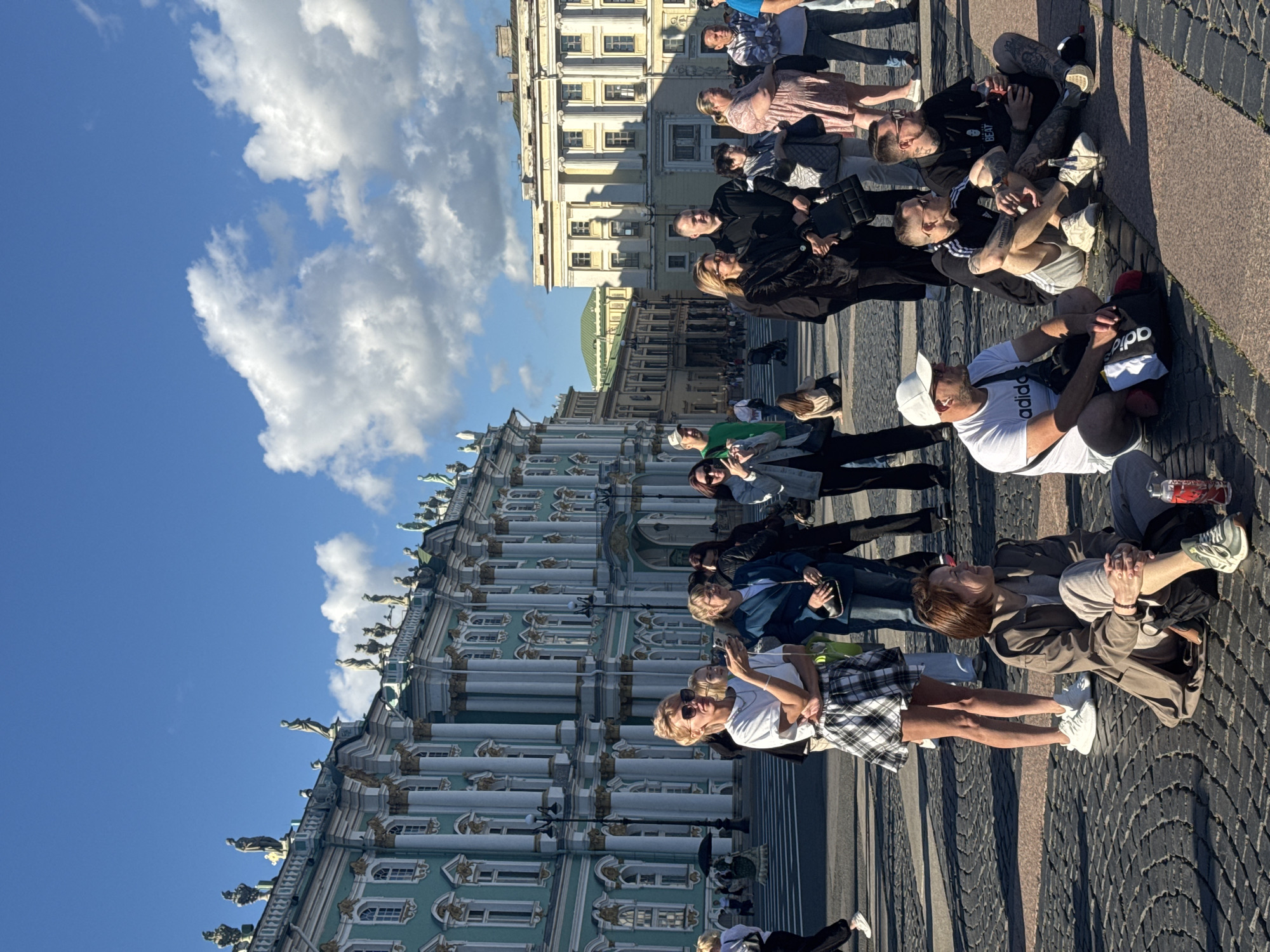 The image captures a sunny day in Palace Square, Saint Petersburg, Russia, a vast open space paved with cobblestones. The iconic green and white facade of the Winter Palace, a grand Baroque building adorned with sculptures and classical columns, dominates the background. Several groups of people are present in the square, some standing and observing, others seated on the ground.
In the foreground, three men are sitting on the cobblestones. The man on the left wears a white t-shirt with "adidas" printed on it and a white baseball cap. The middle man has tattoos on his arms and legs and is wearing a black t-shirt and grey shorts. The man on the right, also tattooed, is wearing a black t-shirt and black shorts, holding a bottle of soda.
Further back, a woman in a trench coat and light-colored pants is sitting on the ground, looking to her right and smiling. To her left, a group of people are standing. A woman with blonde hair, wearing a white t-shirt and a plaid skirt, is holding a phone. A woman in a blue blazer and a black purse stands next to her.
Other individuals and small groups are scattered throughout the square, some appearing to be tourists or locals enjoying the public space. The sky is a clear blue with scattered white clouds. The overall scene depicts a lively and pleasant atmosphere in a historically significant location.