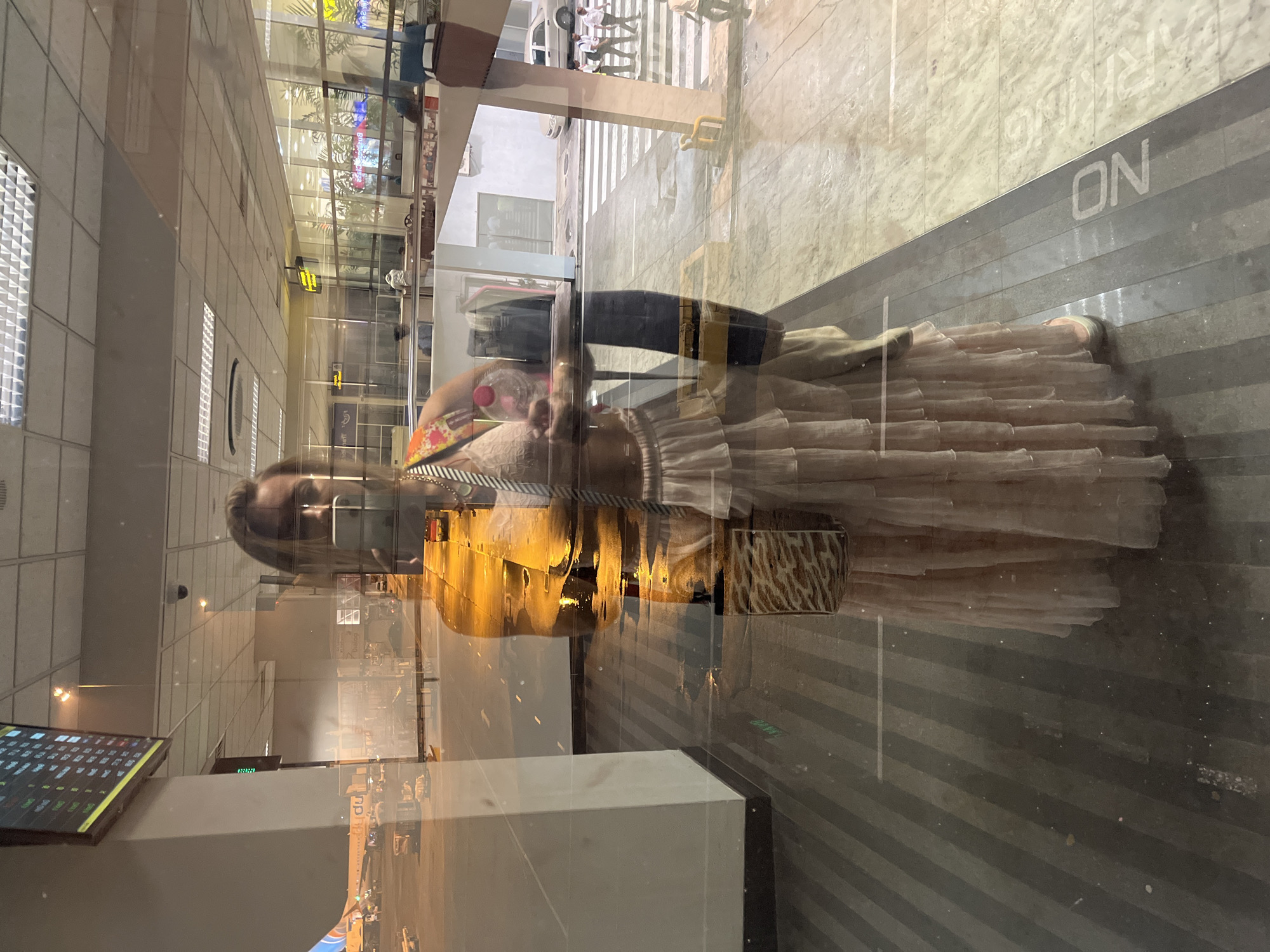 This media file shows a reflection of a woman standing in what appears to be an indoor, modern setting with large glass windows. The woman is wearing a light-colored, ruffled dress and a mask, and is holding a pink object and a bottle. She is looking towards the camera, and her reflection is visible in the glass.
The scene outside the glass appears to be a parking area with striped flooring and parked cars, suggesting a public space such as an airport or a shopping mall. There is also a sign visible in the reflection that reads "PARKING ON". The lighting suggests it is either dusk or nighttime.
The image's orientation is rotated, with the subjects appearing vertically. The overall atmosphere is a bit distorted due to the reflection, creating a sense of depth and layering. The environment seems clean and well-lit, with a mix of natural and artificial light sources.
The provided location context is Katunayake, Sri Lanka.