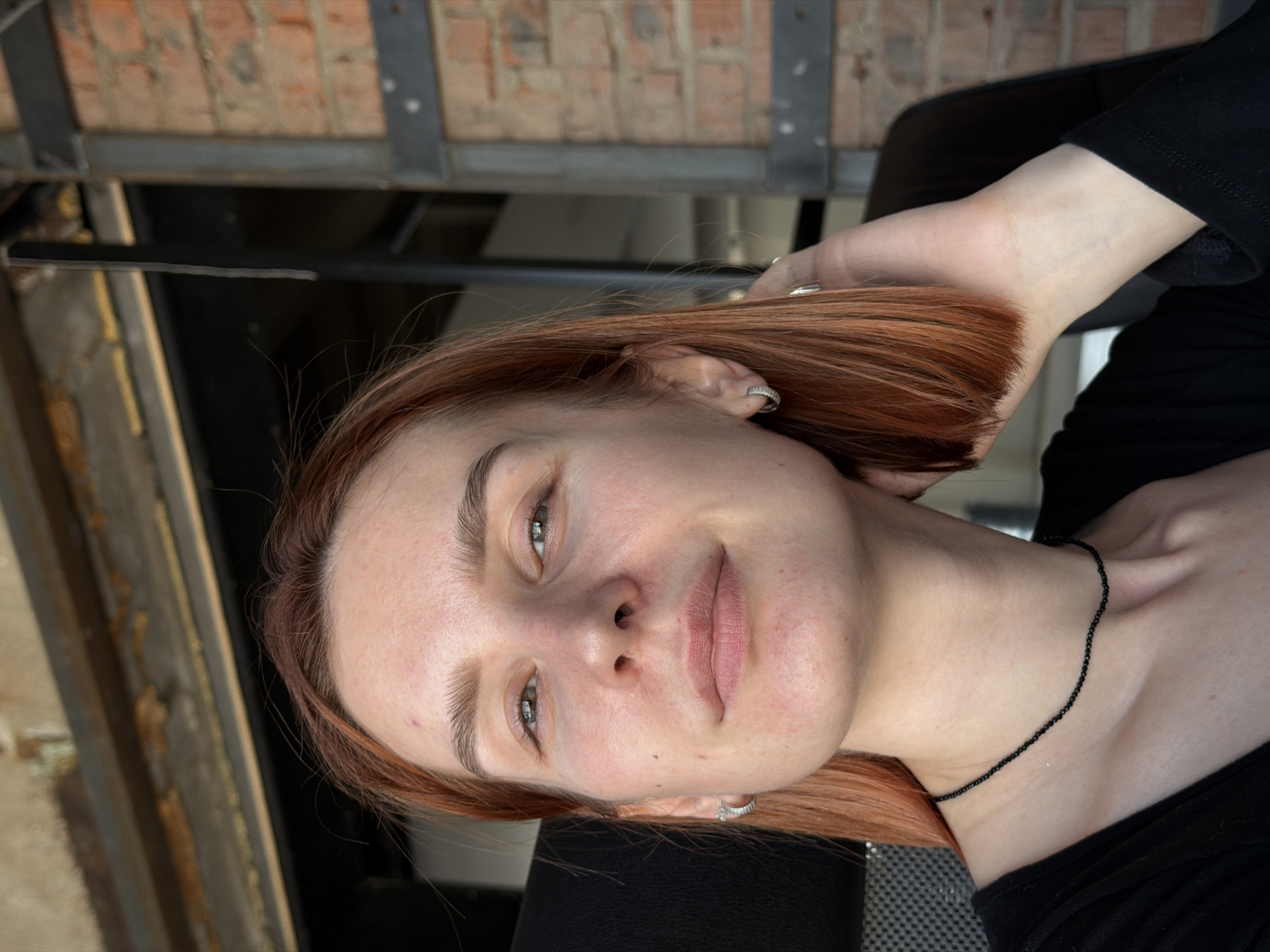 A medium shot depicts a young woman with light skin and reddish-brown hair, wearing a black top and a delicate black beaded necklace. She has light green eyes and no visible makeup. She is smiling softly. Her right hand is raised slightly, positioned near her head.
The background is a mix of interior architectural elements. In the top left corner, there's a partial view of what appears to be a brick structure supported by metal beams. Directly behind the woman, dark shadows obscure details, suggesting a deeper interior space. The surface underneath the woman is a gray material with a textured pattern, potentially a chair or seating.
Although the prompt requests the location, it is not possible to determine the location as Chaikovsky, Russia based on the visual information provided in the image.