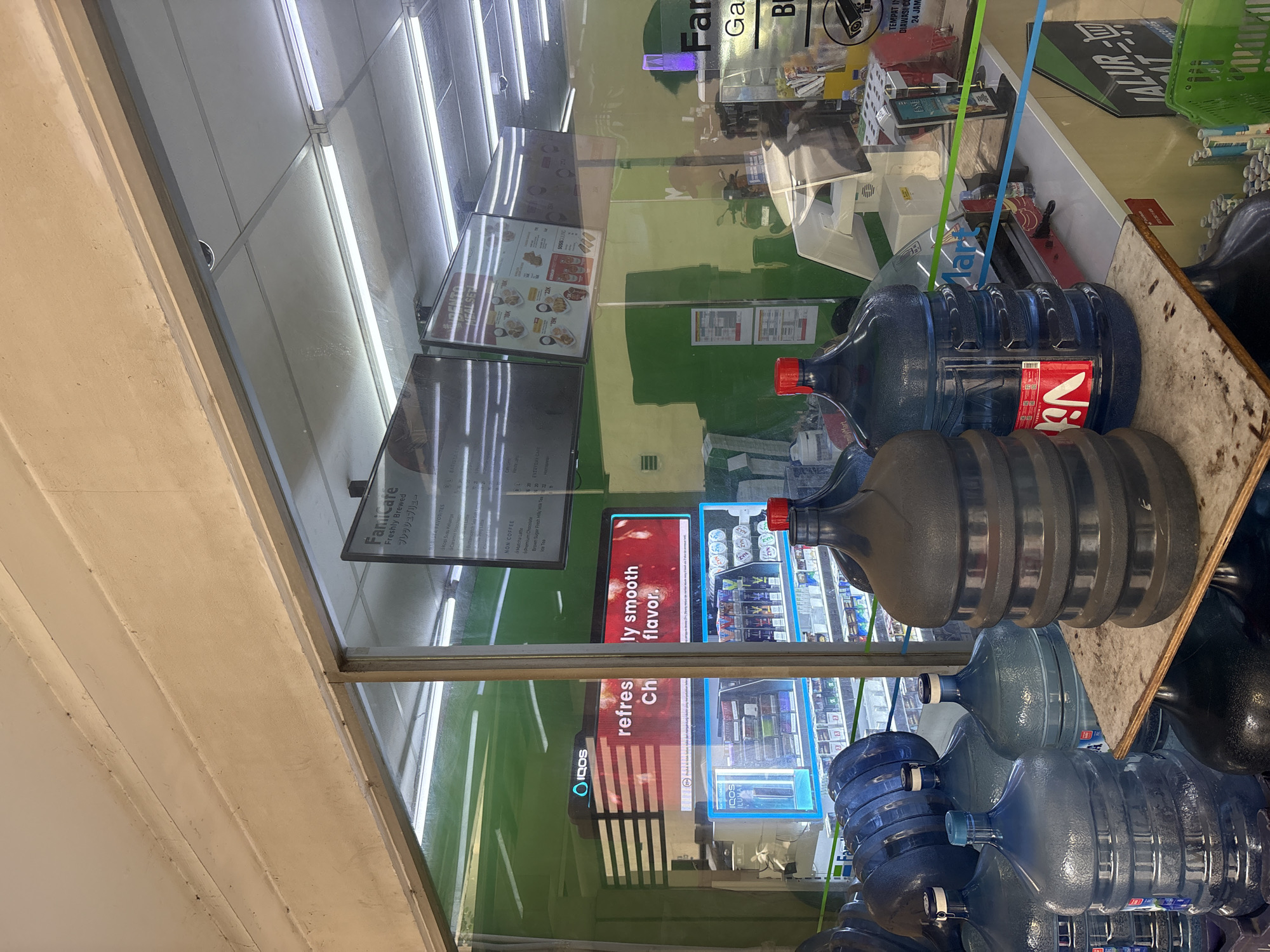 The image shows a collection of large, clear and dark blue plastic water jugs stacked and placed in front of a convenience store window. The store appears to be a FamilyMart, as indicated by a partial sign visible on the right. Inside the store, electronic displays show menu items and promotions, including a "FamiCafe" sign. There is also a refrigerated display with products, and what looks like an IQOS electronic cigarette display. The overall environment suggests an indoor commercial space, likely a convenience store or small supermarket, with a focus on beverages and convenience items. The water jugs are the most prominent objects in the foreground, some stacked high, with red and blue caps. The reflection on the glass window distorts some of the interior details.