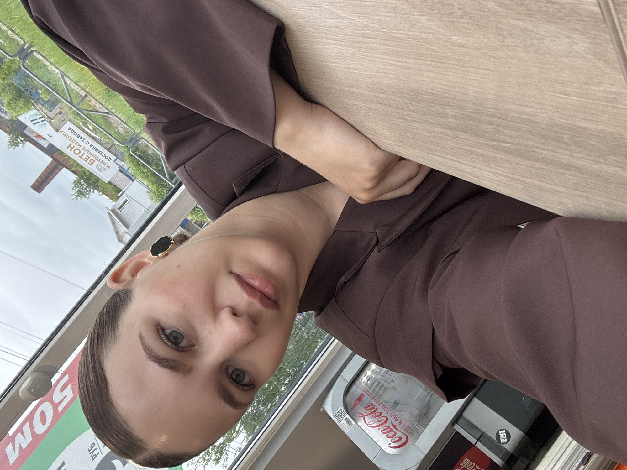 In a selfie taken from a slightly high angle, a young woman with her hair pulled back is looking directly at the camera. She is wearing a brown jacket and a dark earring in her visible ear. Her left hand is resting on a light-colored wooden surface. In the background, through a window, a street scene in Chaikovsky, Russia, is visible, including a building with a chimney and a billboard advertising "BETOH И БЕТОННЫЕ ИЗДЕЛИЯ" (Concrete and Concrete Products), with "ДОСТАВКА С ЗАВОДА" (Delivery from the Factory) below it. A Coca-Cola dispenser is also partially visible on the left side of the frame. The sky is overcast.