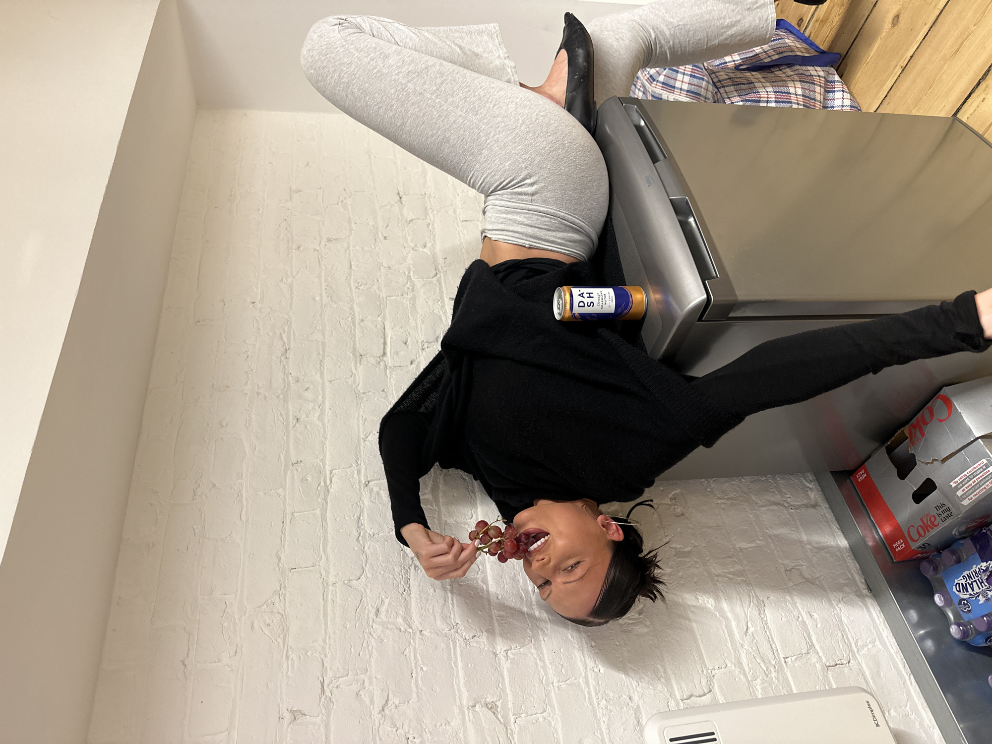 The image depicts an interior scene, presumed to be within an unspecified private residence in the city of London, United Kingdom. A female individual, appearing to be a young adult with dark hair pulled back, is positioned horizontally. Her mid-back rests on the top surface of a metallic Beko brand compact refrigerator or freezer unit. She is wearing a black long-sleeved top and light grey flared-leg trousers, with black flat shoes on her feet. Her upper body and head extend downwards towards the left, with her head tilted to her right, mouth open in an expressive manner, exposing teeth. Her right hand holds a small bunch of red grapes near her open mouth. Her left arm hangs downwards, out of frame at the elbow. A blue and orange can of DASH Orange Sparkling Water is placed upright on the top surface of the appliance, near her hip.
The background consists of a white-painted brick wall with visible mortar lines. To the lower left, a white Dimplex electric panel heater is mounted on the wall. Below this, on a metallic surface, are a partially crushed red Coca-Cola "MEGA PACK" cardboard box and a pack of Highland Spring bottled water. The floor on the right side of the image is composed of light brown wooden floorboards. A blue, white, and red plaid patterned reusable shopping bag rests on the floor adjacent to the refrigerator. A white ceiling and a visible white overhead beam are present at the top of the frame.