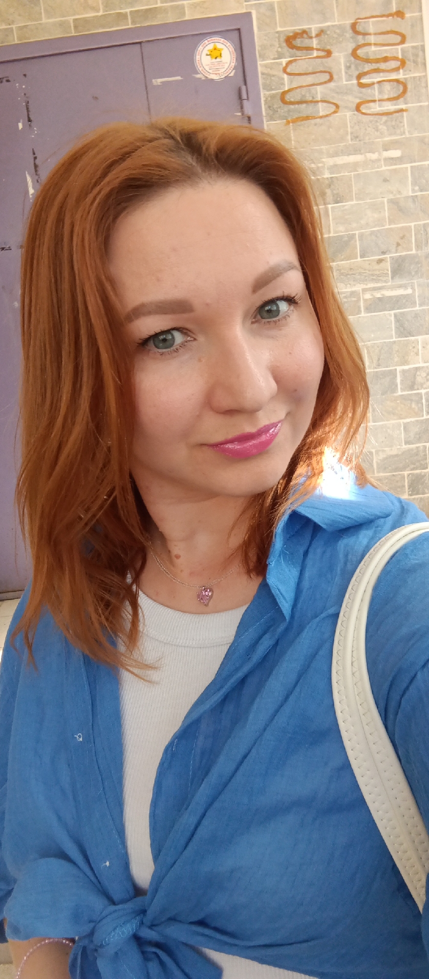 A fair-skinned, auburn-haired woman looks directly at the camera with a slight smile in an outdoor setting. She has green eyes, pink lipstick, and is wearing a delicate silver necklace with a pink pendant. She is dressed in a white ribbed tank top underneath a blue linen button-down shirt, casually tied at the waist. A white shoulder strap of what appears to be a bag is visible. The background consists of a lavender-colored door with a round sticker near the top, as well as a textured brick wall with two abstract brown squiggles painted on it. Location: Unknown, Unknown.