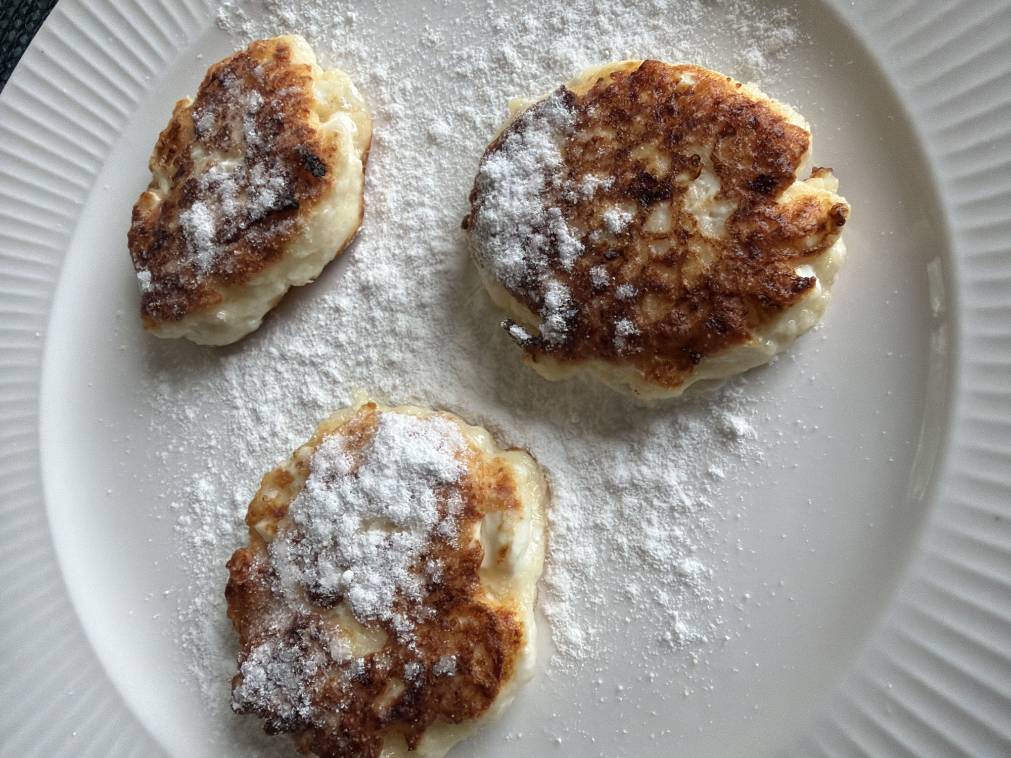 Three freshly prepared, pan-fried fritters, likely traditional Russian syrniki (cottage cheese pancakes), are prominently displayed on a white ceramic plate. Each of the three fritters is roundish with a slightly irregular shape, featuring golden-brown, crispy surfaces and lighter, softer centers, indicative of having been pan-fried. They are generously dusted with white powdered sugar, which also scatters across the white surface of the plate, creating a delicate, snowy appearance.
The plate itself is white ceramic and has a distinctive ridged pattern along its outer edge, providing a subtle textural detail to the clean background. The composition is a close-up shot, focusing entirely on the food, presented as if ready to be eaten. The soft, even lighting suggests an indoor setting, possibly for breakfast or brunch. The image conveys a sense of homemade comfort food, a common culinary scene in locations like Vaskelovo, Russia, where syrniki are a popular dish. There is no visible text in the image.