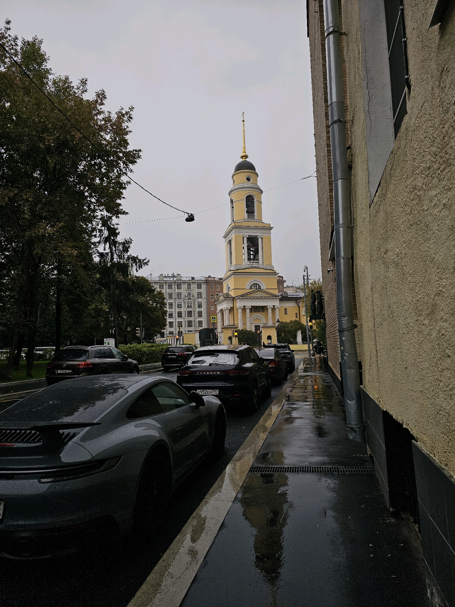 The image shows a street scene in Moscow, Russia, on an overcast day, with the pavement wet from rain. A gray Porsche is parked in the foreground on the right side of the street, facing away from the camera. Behind it, a line of other cars, including dark SUVs, are parked and in motion. A tall, yellow church with a gold spire and bell tower dominates the background on the left side of the street. The church is flanked by buildings with classical architecture. Trees line the left side of the street, and a textured, yellow building with a gray drainpipe runs along the right side of the image. The wet road reflects the surrounding lights and the sky. There are no visible people in the immediate foreground or midground.
