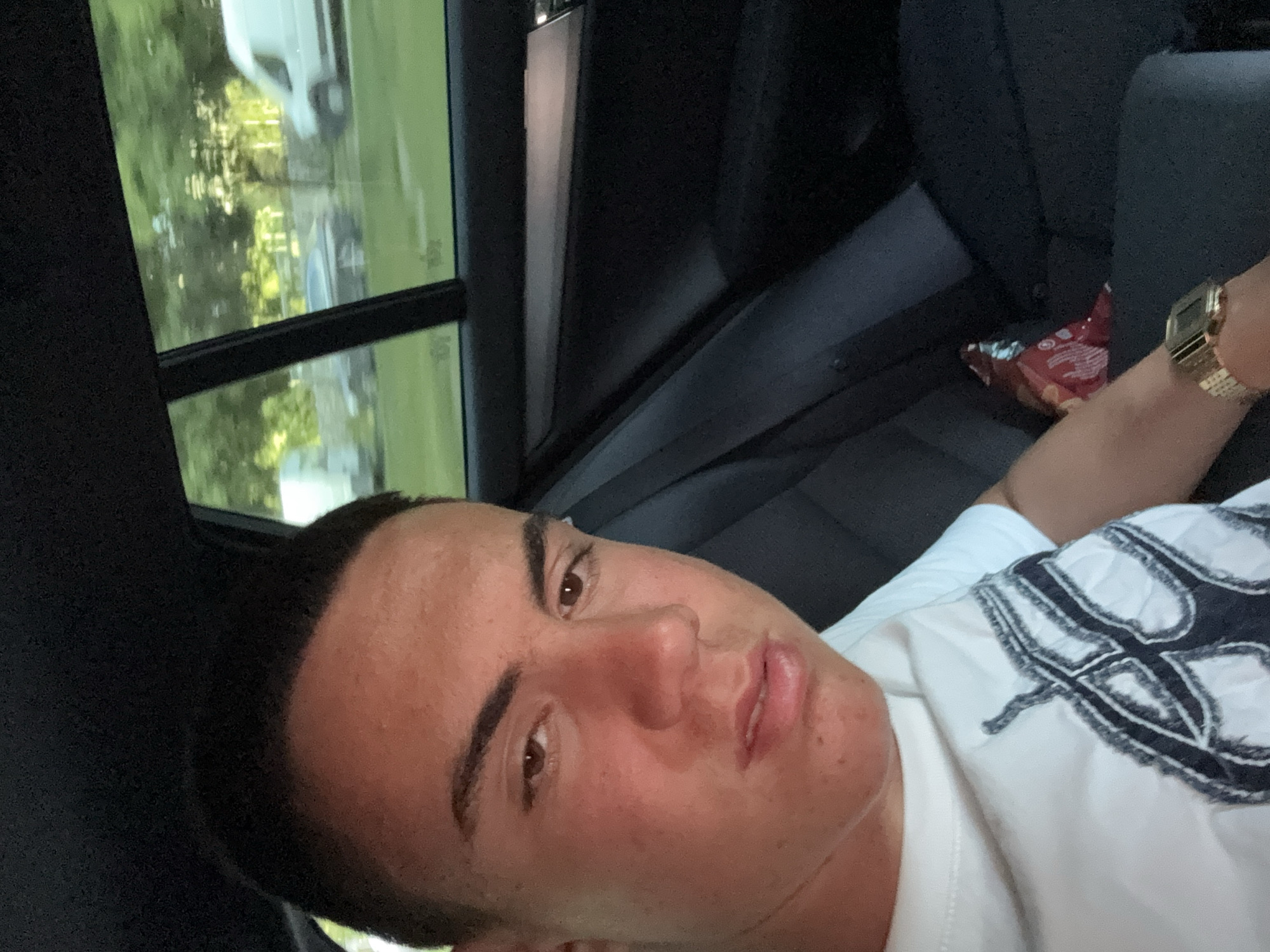A close-up, slightly rotated image of a young man with dark hair, light skin, and brown eyes. He is wearing a white t-shirt with a dark embroidered design. He appears to be sitting in the back seat of a car, based on the seat fabric, window, and the presence of an open bag of potato chips and a gold wristwatch on his arm. The view outside the window includes greenery and a glimpse of a white vehicle. The location is indeterminate; there is no indication this is Plovdiv, Bulgaria.