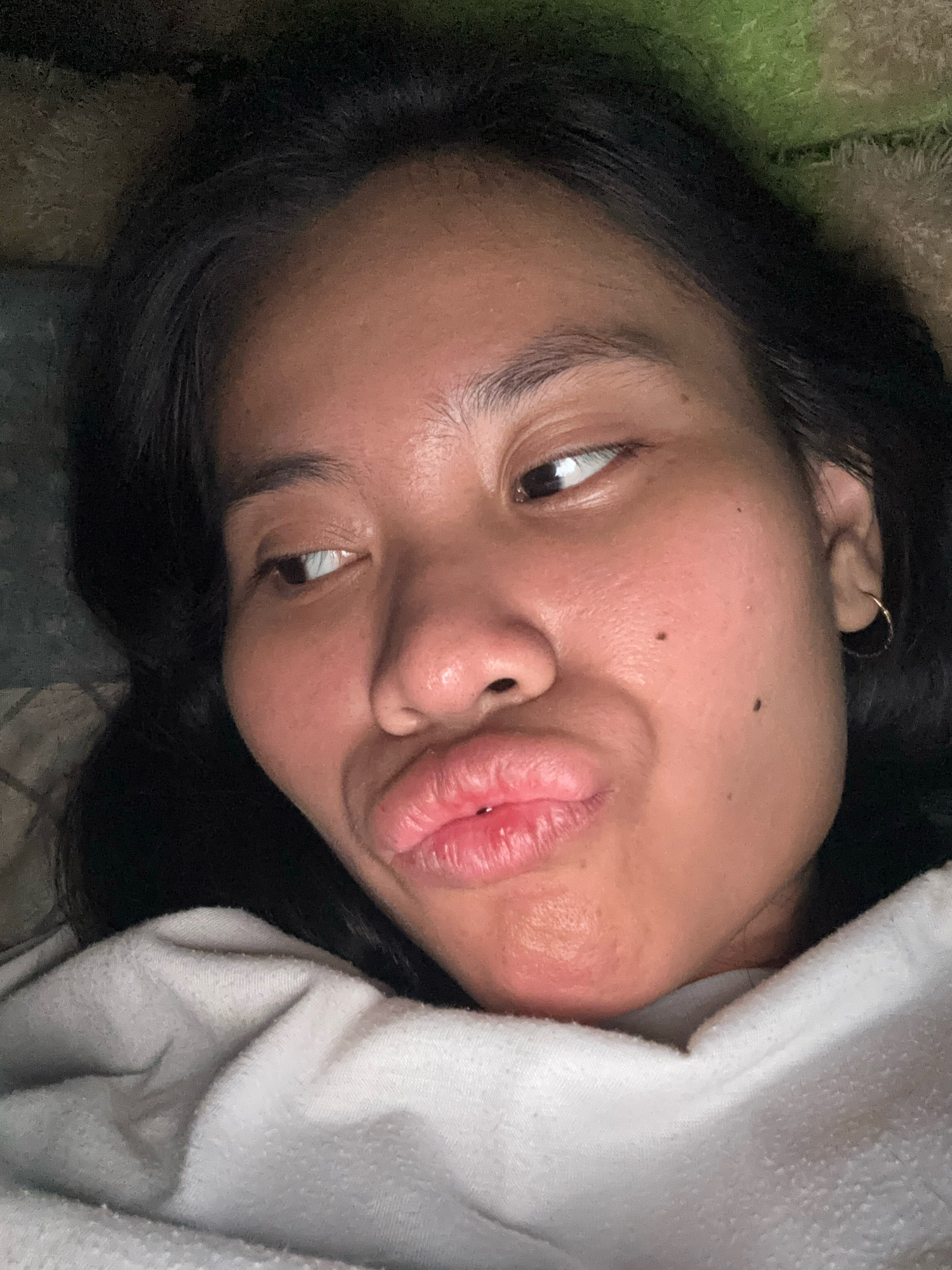 The image is a close-up selfie of a young woman with dark hair, who is lying down and making a kissing face. She is wearing a light-colored hooded garment. The background appears to be a bed with patterned sheets and a green textured surface. There are no visible indicators to suggest this image was taken in East Jakarta, Indonesia.