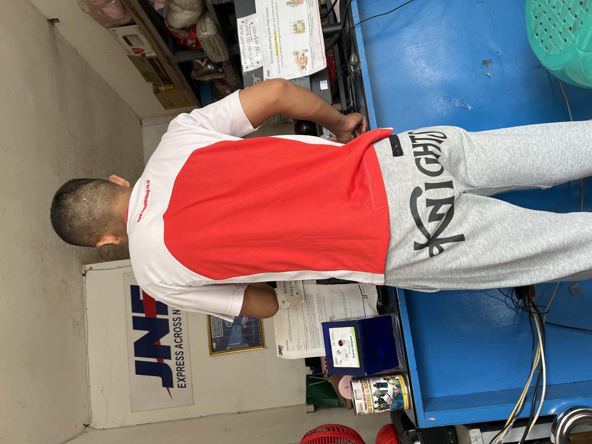 The image depicts an indoor scene, likely a small business or service point, in Citeureup, Indonesia. The focal point is a young man standing with his back to the camera, wearing a white t-shirt with a large red panel on the back and grey sweatpants with the word "KNIGHTS" printed in black. He is positioned behind a blue counter.
Behind the man, a white sign with the JNE logo and "EXPRESS ACROSS N" is mounted on the wall. Several documents and posters are visible on the counter and wall, including a JNE shipping form and a notice about peak season shipping. To the left of the counter, a red electric fan is partially visible, along with a cylindrical container holding various items and a small blue dispenser with a sticker showing two children. Shelving units filled with various items are present in the background, with some items wrapped in plastic. The overall environment appears somewhat cluttered but functional, characteristic of a local service establishment.