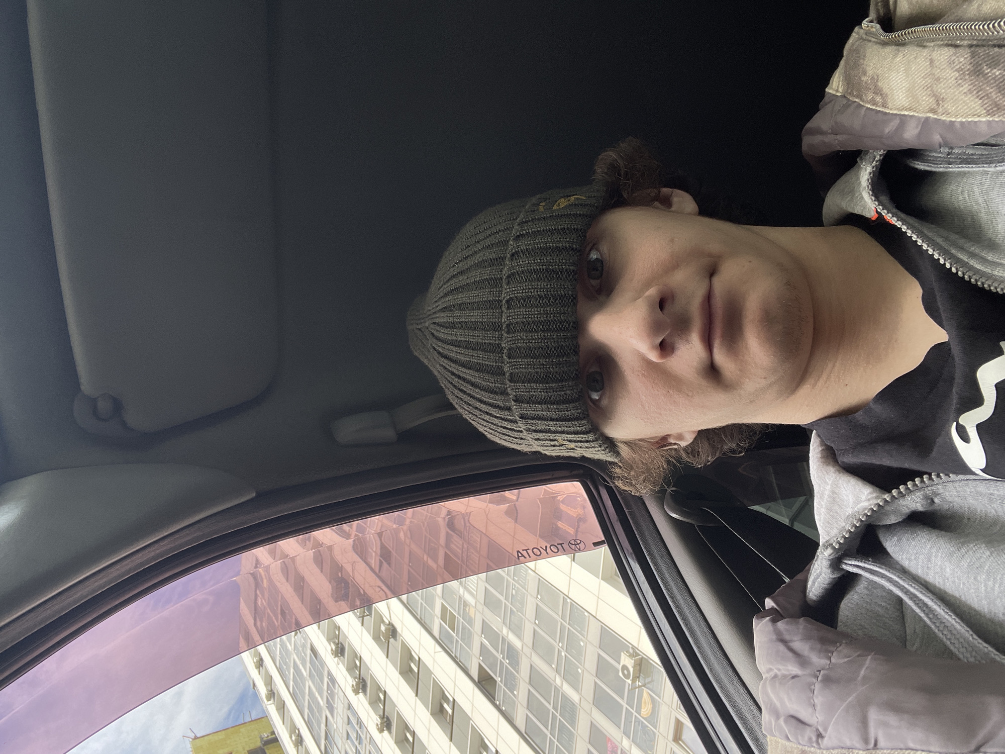 A person wearing a gray beanie, black shirt, and gray jacket is visible inside a car. The window has a pink tint and displays "AToyOT," likely a misspelling or stylized representation of the brand "Toyota." Outside the car window is a tall, multi-story building with a grid-like pattern of windows, suggesting an apartment complex or office building. The location is Krasnoyarsk, Russia, based on the lettering in the Toyota logo. The person appears to be taking a selfie or picture from inside the vehicle.