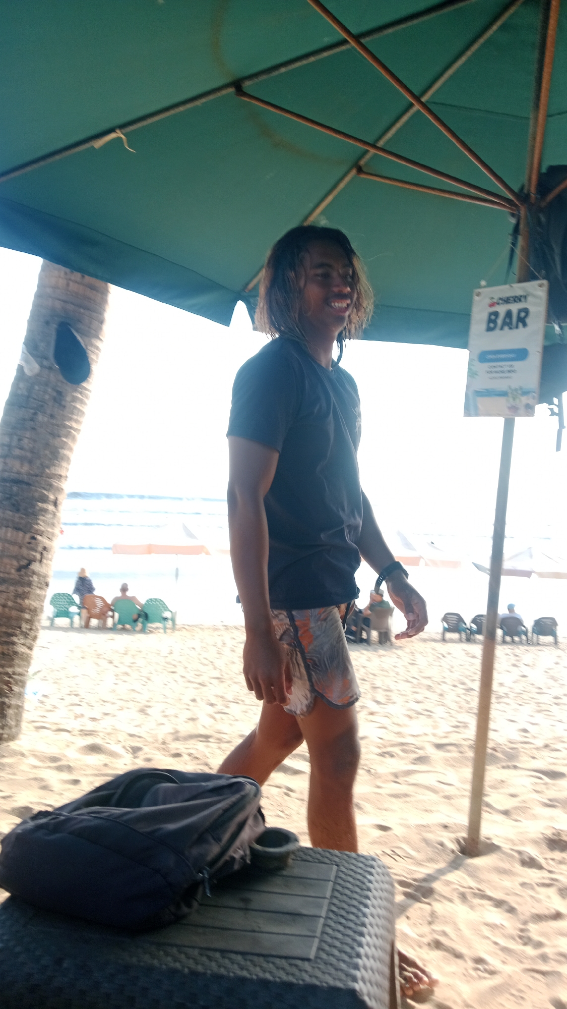 A young man with shoulder-length, dark, wavy hair walks on a sandy beach in Kuta, Indonesia, under a large green umbrella. He is wearing a dark t-shirt and patterned swim trunks. He is smiling and looking to his right. In the foreground, a black backpack rests on a woven table. In the background, the ocean is visible with gentle waves, and several beach chairs are set up on the sand. A sign with the word "BAR" is mounted on a pole near the man. Palm trees are also present on the beach. The lighting suggests it is daytime, with bright sunlight.