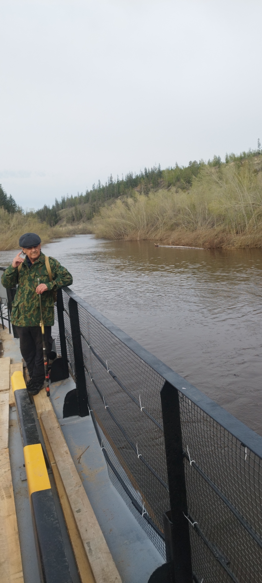 A full-length shot depicts an elderly man standing on a river ferry, likely in Mokhsogollokh, Russia, given the description. The man is wearing a camouflage jacket, dark pants, and a flat cap. He is holding a fishing rod and appears to be looking out over the river. The ferry has a dark-colored metal railing with a wire mesh, and wooden planks are visible on the deck. The river is a muddy brown color, and the far bank is lined with trees and vegetation. The sky is overcast.