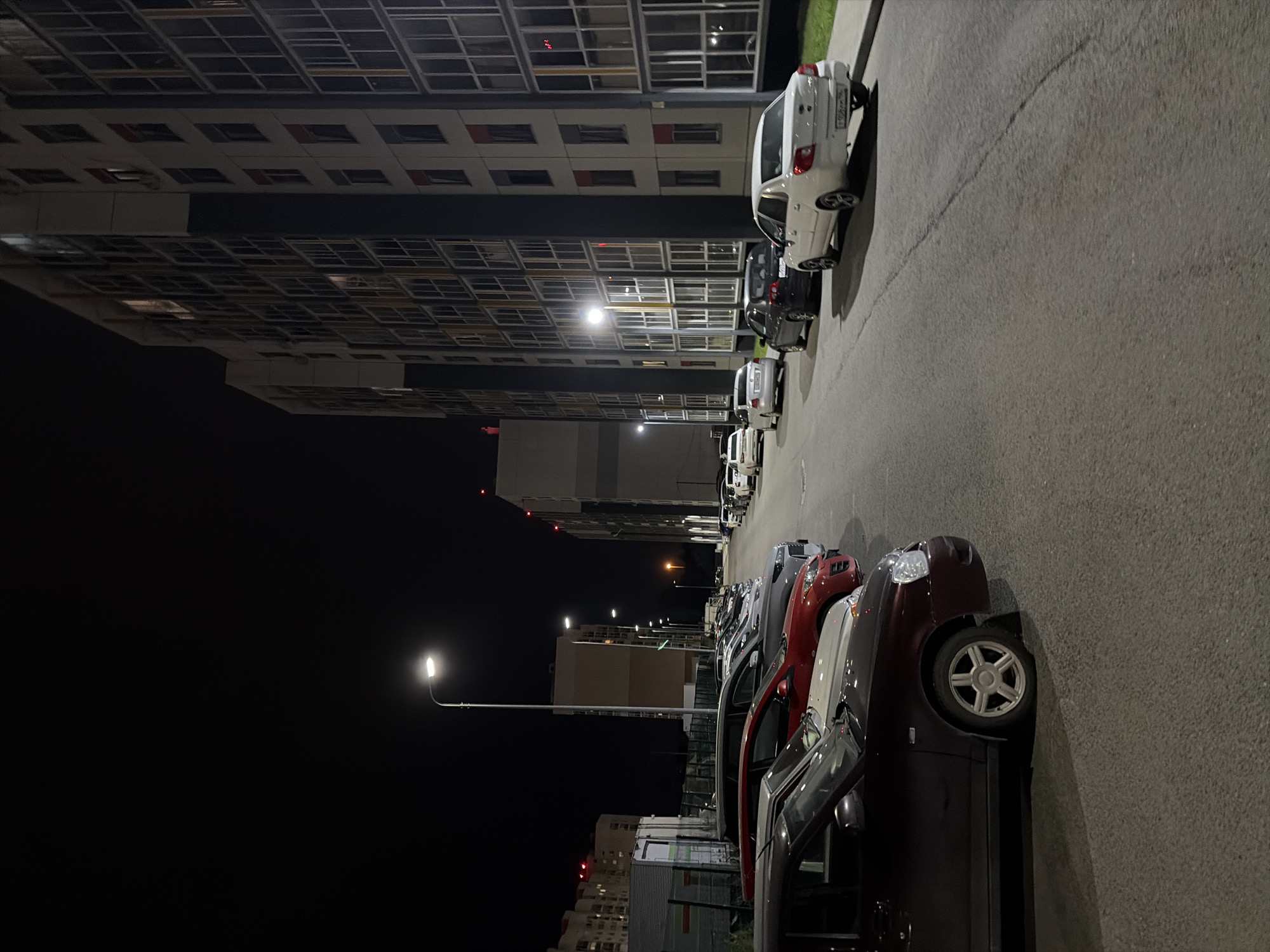 The image depicts a nighttime scene in a residential area of Yekaterinburg, Russia. A paved road runs alongside a modern, multi-story apartment building with numerous windows illuminated from within. Several cars are parked on the side of the road, aligned parallel to the building and the curb. Streetlights cast a glow on the road and vehicles. The sky is dark, indicating it is nighttime. The overall atmosphere is quiet and still.