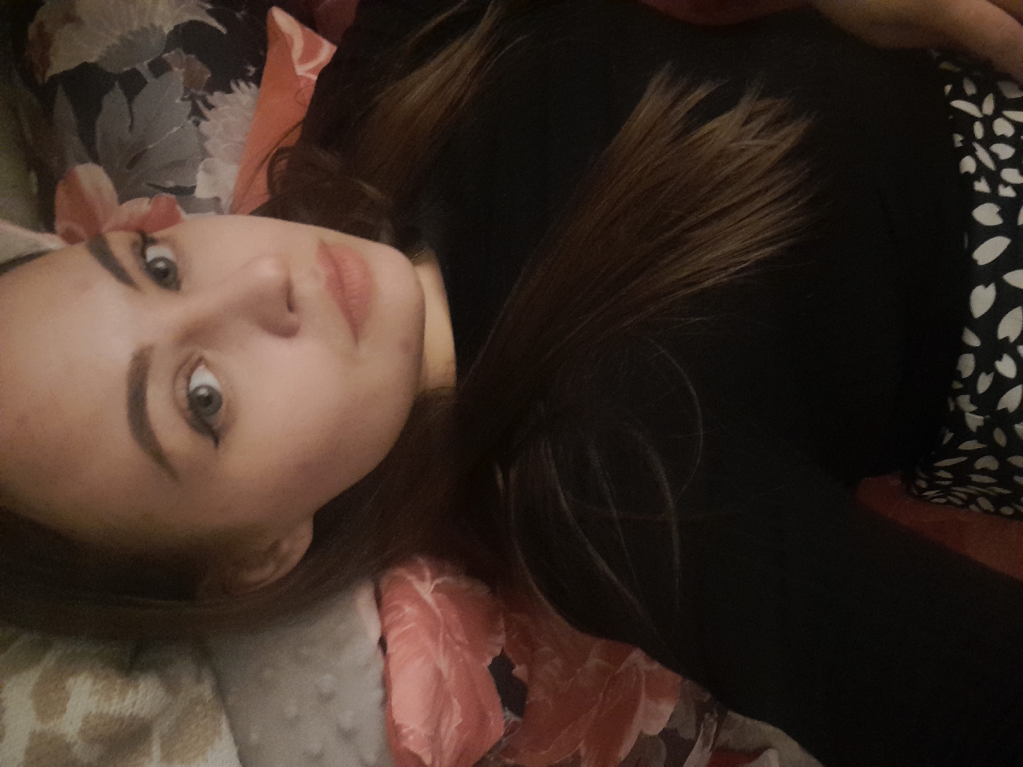 A woman is lying down, facing upwards towards the camera. She is wearing a black long-sleeved shirt. Her hair is brown, and her face is pale with defined eyebrows and eyeliner. The background includes a patterned bedspread featuring floral designs and polka dots. The location is Unknown, Unknown.