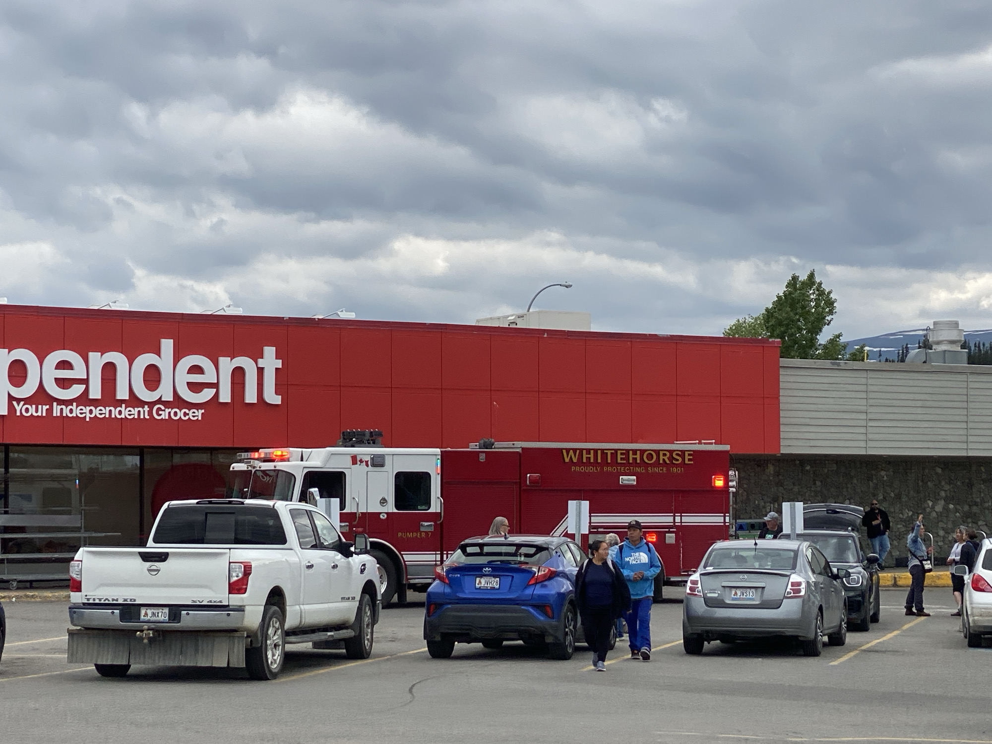An overcast sky looms over a parking lot in front of a large red building with "Independent Your Independent Grocer" written on it in large white letters. A white pickup truck is parked in the foreground. Behind it, a fire truck with "WHITEHORSE PROUDLY PROTECTING SINCE 1901" is visible, along with a blue sedan. Several people are walking in the parking lot. The scene appears to be a typical day at a grocery store, with the fire truck possibly a routine presence or responding to a minor incident.