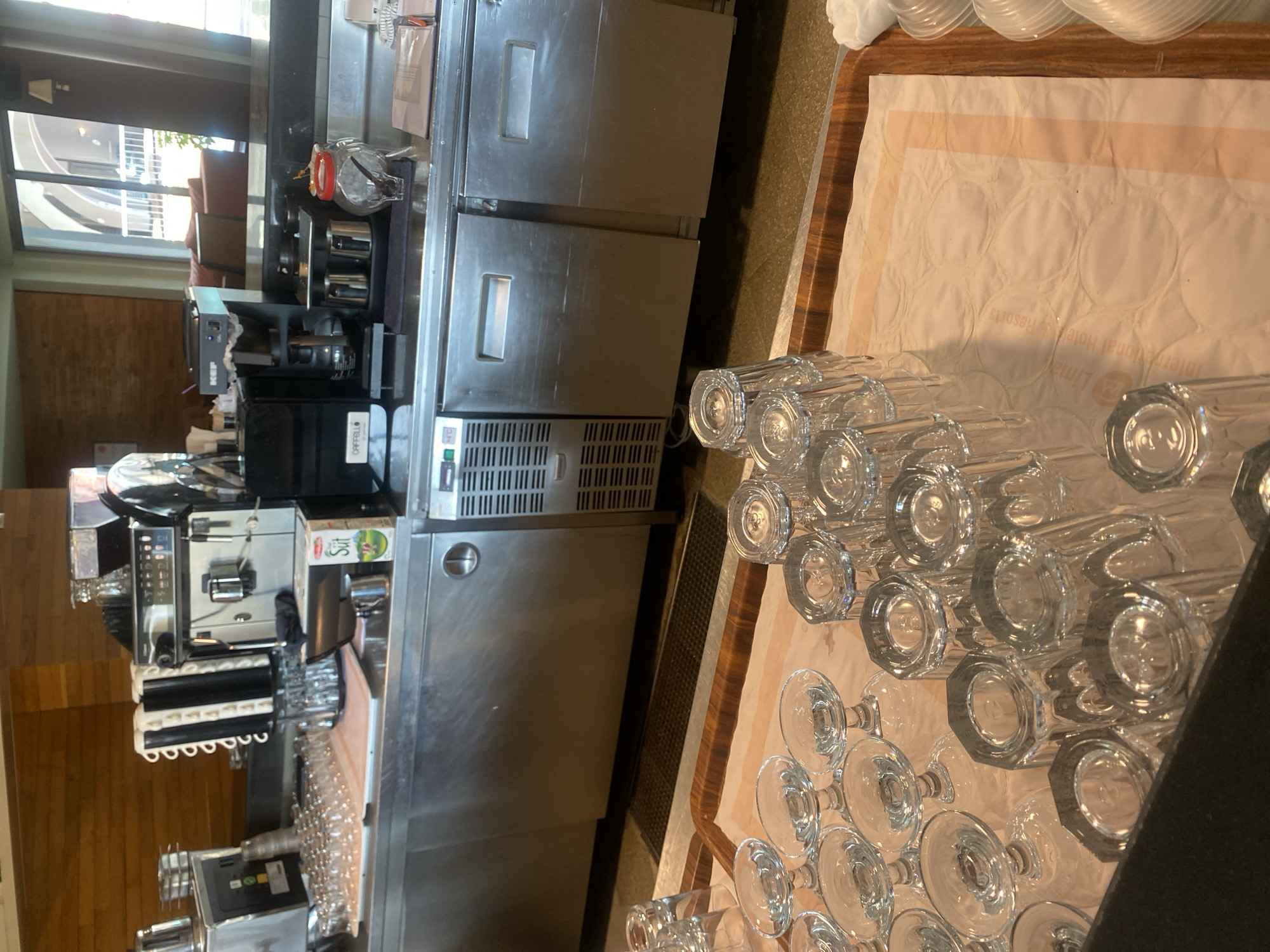 A stainless steel bar counter is laden with various items. In the background, a large coffee machine and a beverage dispenser are visible. The counter also holds several rows of clean drinking glasses, stacked neatly on top of each other. Some glasses are upside down, suggesting they are ready for use. A small carton of milk is placed on the counter. The environment appears to be a commercial establishment, possibly a hotel or restaurant, given the presence of bar equipment and extensive glassware. The overall impression is one of preparedness and organization within a service setting. The location in the city of Aksu, Turkey is not discernible from the image itself.