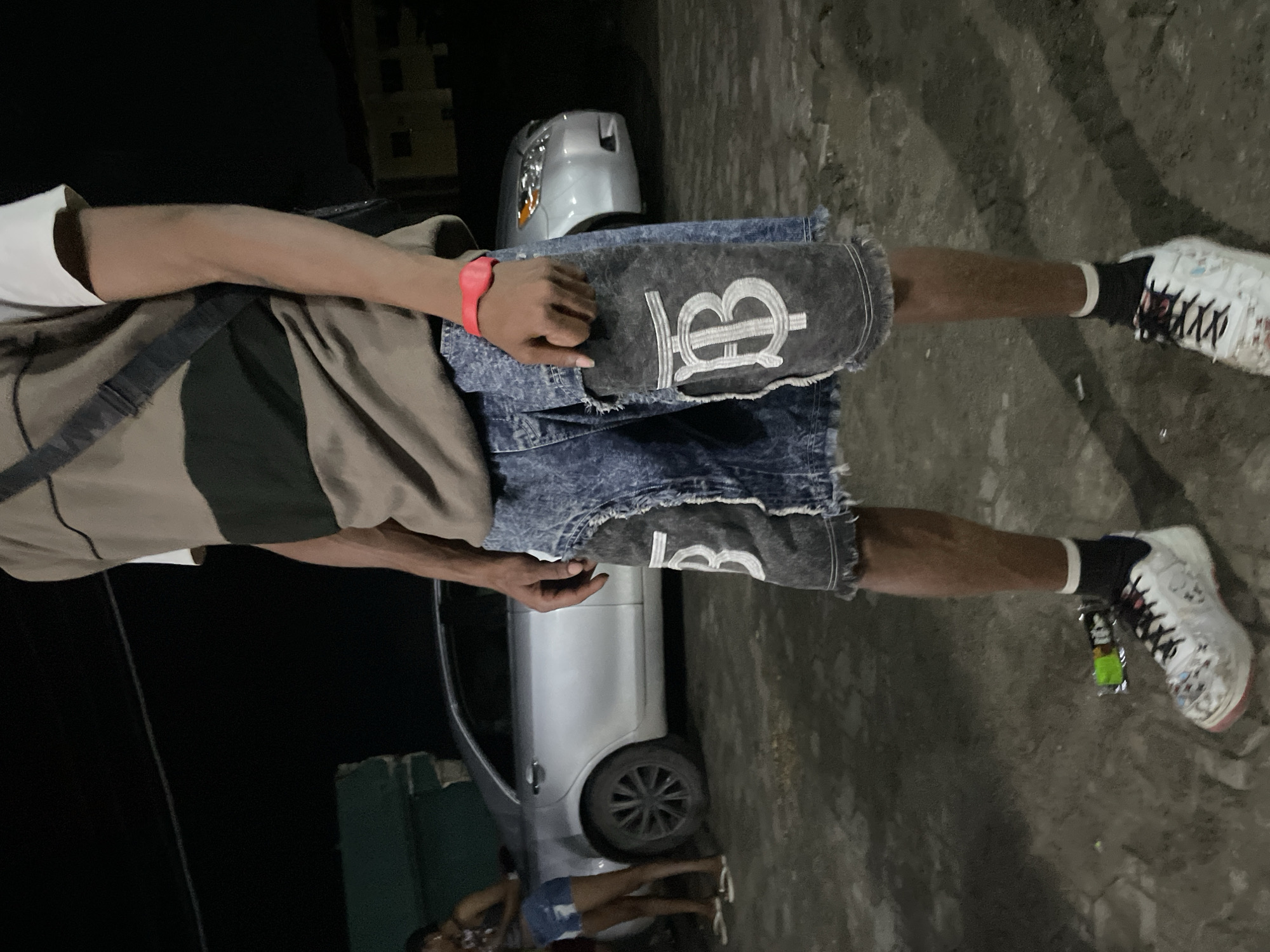 The image depicts a nighttime scene in Ado, Nigeria. In the foreground, a person is shown from the waist down, wearing denim shorts with distressed patches featuring a white "TB" logo, a brown and dark green striped t-shirt, black socks, and white sneakers. A red wristband is visible on their right wrist. Behind this individual, a silver car is parked on a paved surface. To the left of the car, another person, a woman, is standing and looking down. The background is dark, suggesting it is nighttime, with the faint outlines of buildings visible in the distance. A green, possibly makeshift structure, is also present on the left side of the image. The ground appears to be dirt and gravel.