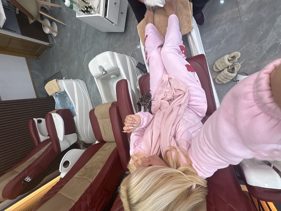 An indoor scene within a nail salon or spa, specifically in Chalfont St Peter, United Kingdom, depicts a blonde female receiving a pedicure service. The subject is seated in a dark red and tan pedicure chair, wearing a pink long-sleeved top and pink pants with red cherry patterns. Her bare feet rest on a tan towel, where an individual wearing dark clothing and white gloves is actively performing a pedicure. The subject's left hand, displaying light pink manicured nails, is visible in the foreground. Two fluffy beige slippers and a white face mask lie on the gray tiled floor beside her. Three pedicure stations are present, each consisting of a large chair and a white foot basin; one basin contains blue liquid and a towel, while another is empty. The walls feature dark wood paneling and a light-colored slatted section. In the background, a white cabinet holding various manicure tools and bottles, along with another chair and a wooden door, are visible.