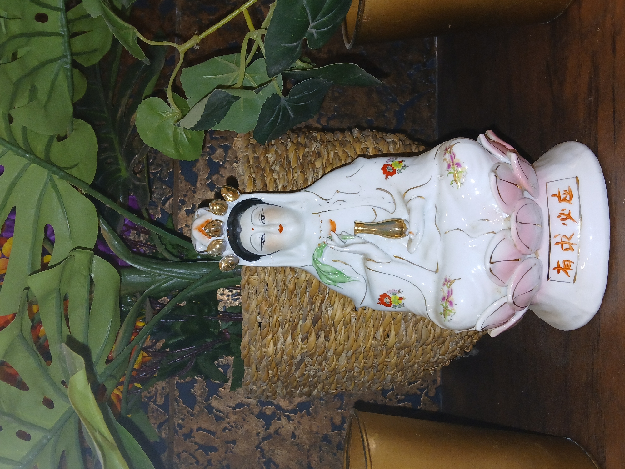 A porcelain statue of a deity is positioned in a basket, which is surrounded by green plants. The statue is predominantly white with floral designs and has golden accents. The basket holding the statue has a woven texture. The background includes a dark, patterned surface. The statue is labeled with Chinese characters written vertically. Bengaluru, India is not visually discernible in the image.