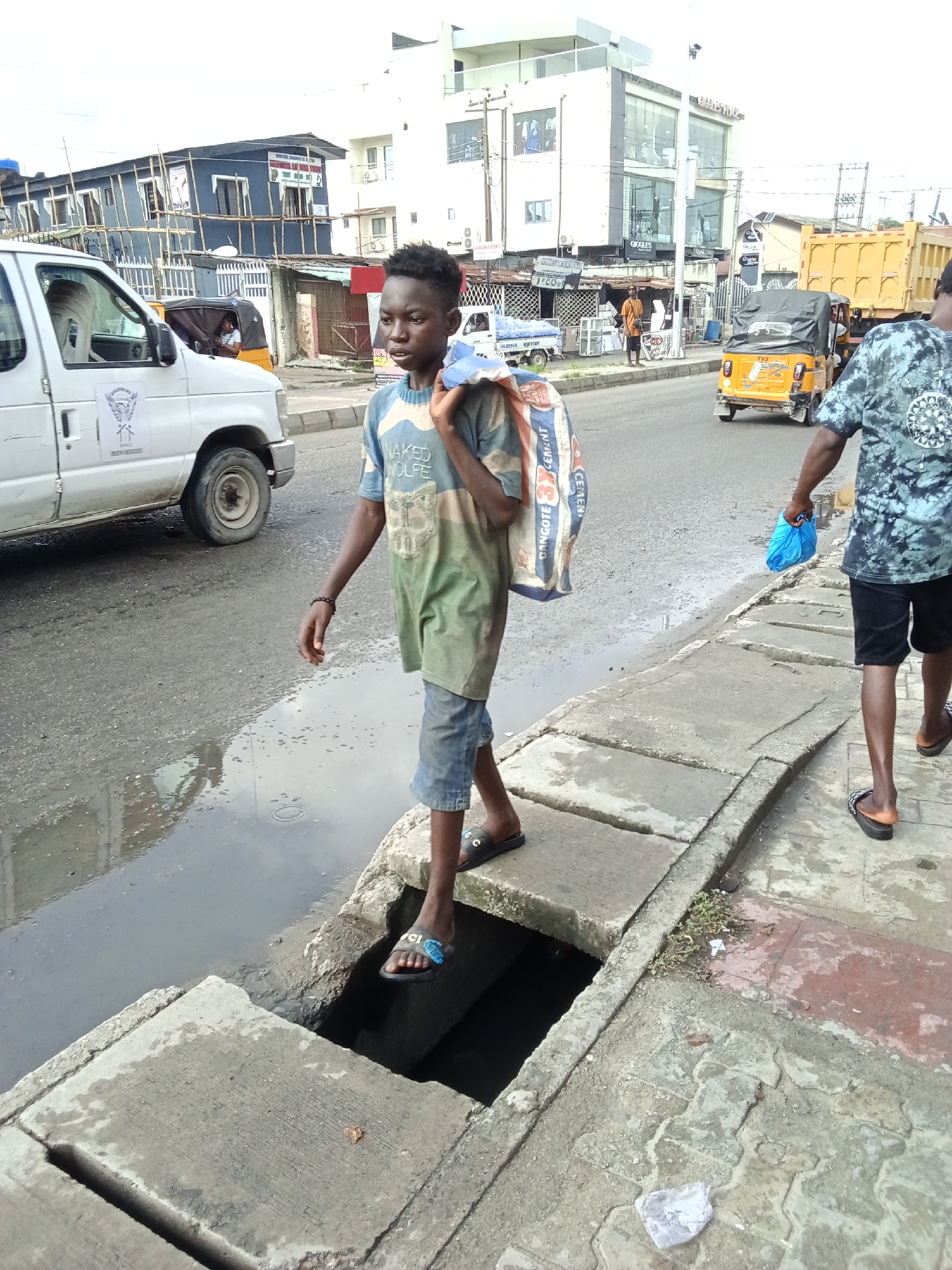 This medium shot captures a bustling urban street scene in Ado, Nigeria, under an overcast sky. In the foreground, a young man walks on a concrete sidewalk, stepping carefully over a section of open drainage ditch filled with dark water. He wears a two-tone t-shirt with "NAKED WOLFE" printed on it, blue denim shorts, and black slides. A large white and blue bag, visibly labeled "DANGOTE CEMENT 3X," is slung over his right shoulder. His gaze is directed towards the left side of the frame.
To his right and slightly behind him, another person, partially obscured, walks on the same sidewalk, carrying a blue plastic bag. The street parallel to the sidewalk is an asphalt road with puddles of water, suggesting recent rain. Vehicular traffic includes a white van with an emblem (possibly an eagle or bird) on its side, a yellow TVS brand auto-rickshaw (keke napep) with a black canopy, and a large yellow dump truck further down the road.
The background features a mixture of commercial buildings. On the left, a blue building with bamboo scaffolding shows signs for "VIRTUAL DIAGNOSTIC & CT SCAN" and "LABORATORY." Further right, a multi-story white building with large glass windows and the word "OROGLES" faintly visible adds to the commercial atmosphere. The overall scene depicts a typical daytime, active urban environment in a West African city.