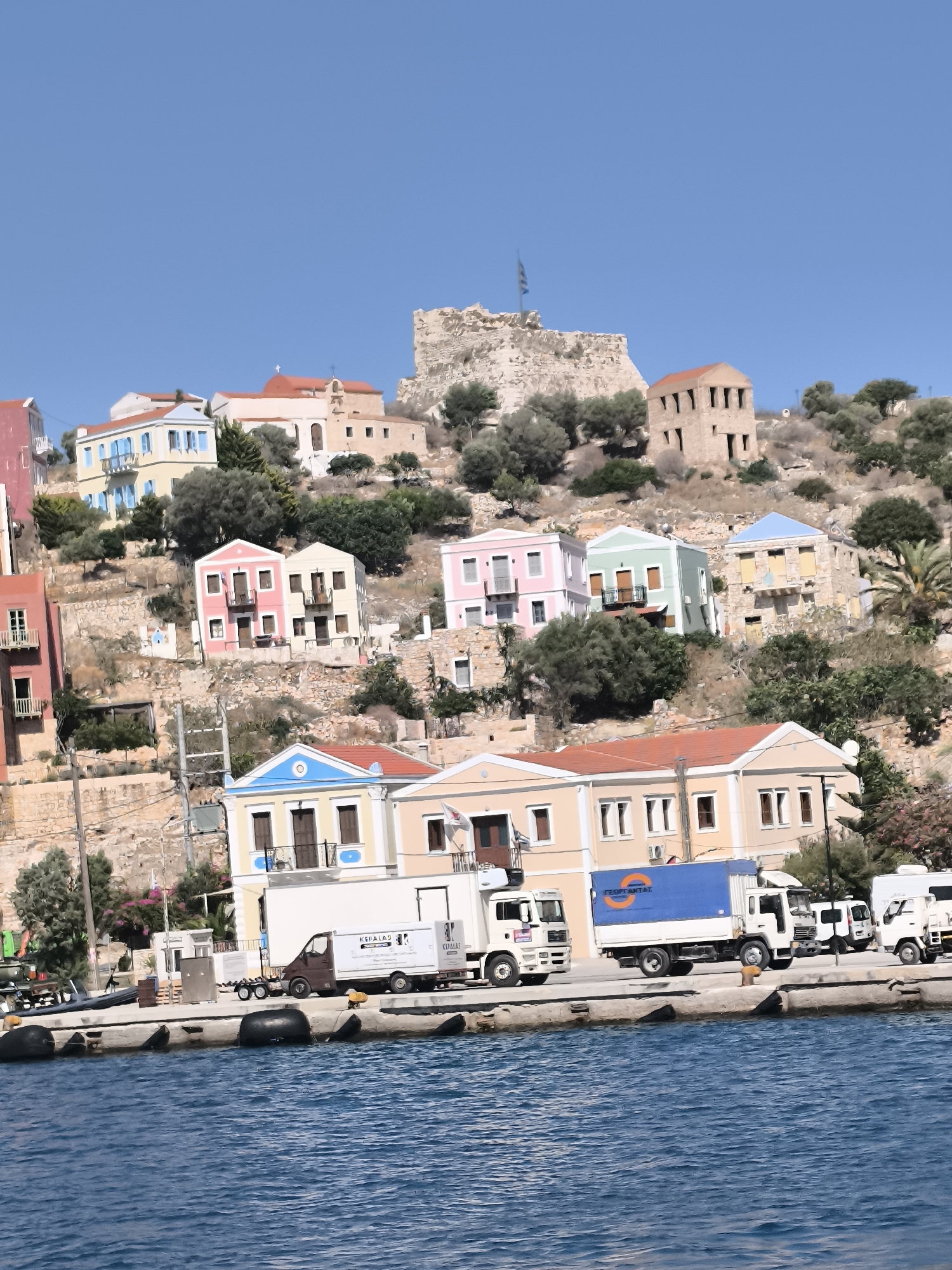 This image captures a waterfront scene in an unidentified location, likely a Greek island or coastal town, characterized by colorful buildings cascading down a hillside under a clear blue sky. In the foreground, the deep blue water of a harbor laps against a quay lined with large black buoys. Several trucks are parked on the quay, including a white truck with a blue container bearing Greek lettering, and a smaller brown truck. Behind the quay, a row of buildings with red-tiled roofs lines the waterfront, with more houses in shades of yellow, pink, and blue built into the steep hillside above. Further up the hill, ancient stone ruins, possibly a castle or fortification, are visible against the sky, topped with a Greek flag. The vegetation appears arid, with olive trees and scrub dotting the landscape. The overall impression is one of a sun-drenched, Mediterranean coastal settlement.