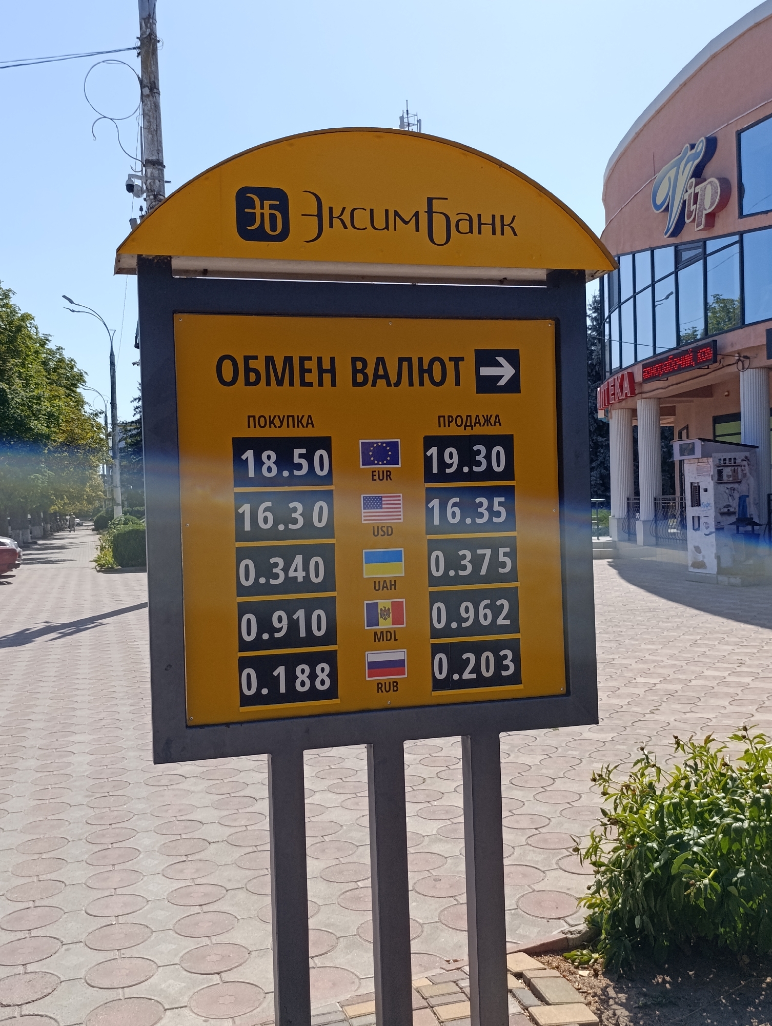 The image displays an outdoor currency exchange rate board in Dubăsari town council, Moldova. The yellow board has black text and numbers indicating exchange rates for different currencies. The top of the board features the logo and name of "ЭКСИМБАНК" (EXIMBANK). Below this, the heading "ОБМЕН ВАЛЮТ" (Currency Exchange) is followed by an arrow pointing to the right.
The board lists five currency pairs: EUR, USD, UAH, MDL, and RUB. For each currency, there are two columns: "ПОКУПКА" (Purchase) and "ПРОДАЖА" (Sale). The purchase rates are listed on the left and the sale rates on the right.
Visible purchase rates are:
EUR: 18.50
USD: 16.30
UAH: 0.340
MDL: 0.910
RUB: 0.188
Visible sale rates are:
EUR: 19.30
USD: 16.35
UAH: 0.375
MDL: 0.962
RUB: 0.203
The flags of the respective countries or currency unions are displayed next to each currency. The background shows a paved walkway with a circular pattern, trees and bushes on the left, and a modern building with a sign that appears to say "TIP" on the right. The sky is clear and blue. There are no people clearly visible in the frame interacting with the board or its immediate surroundings. The overall scene suggests a commercial area.