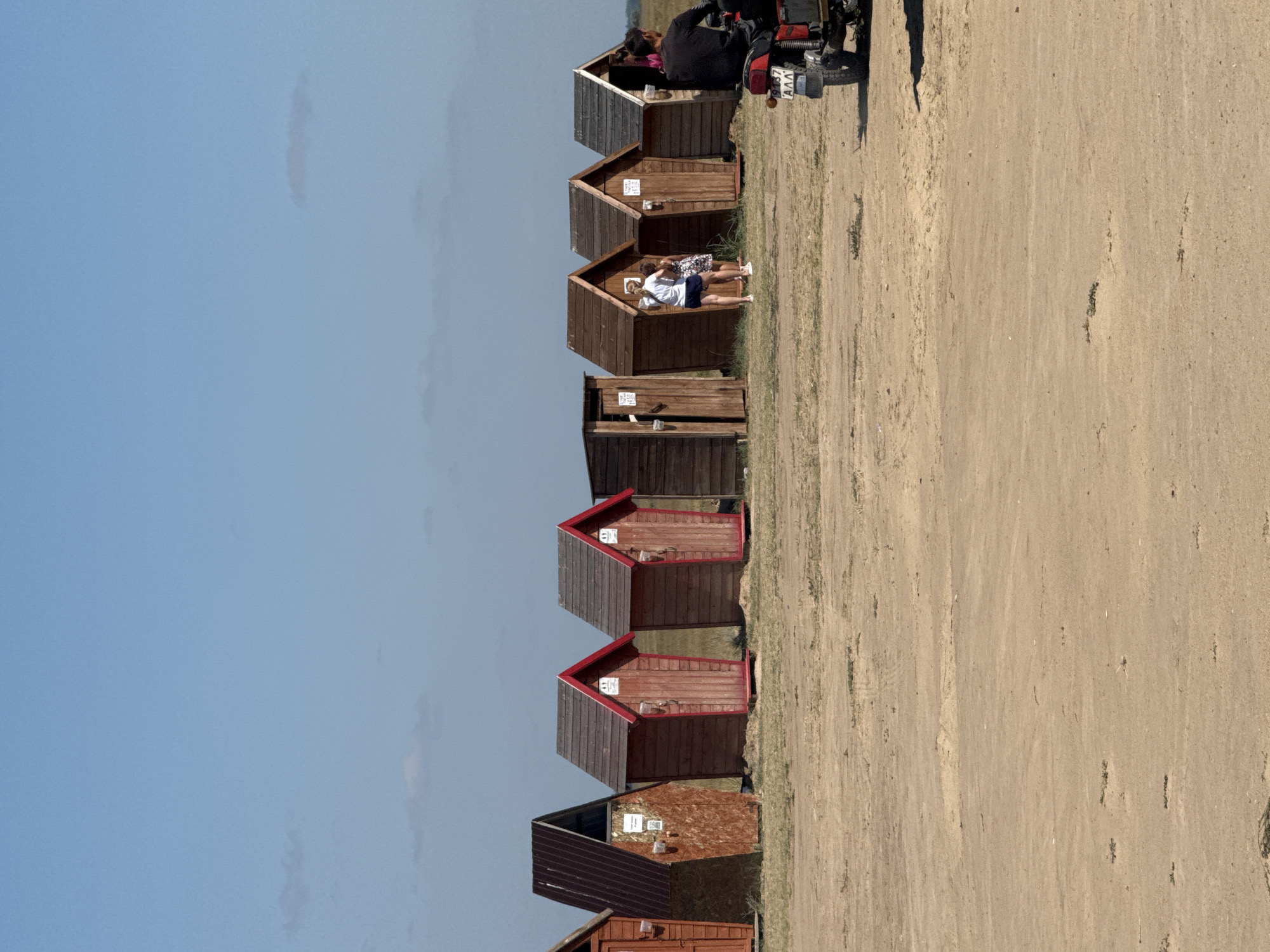 The image depicts a row of small wooden structures, likely changing rooms or outhouses, situated on a sandy or dirt area. The structures have a rustic appearance, with some featuring pitched roofs and wooden doors. The ground is a dry, dusty surface with sparse vegetation. In the background, a clear blue sky is visible, with a few wispy clouds.

To the right of the structures, two individuals are standing near an open doorway. One is a woman wearing a white t-shirt and dark shorts, and the other is a woman in a patterned dress. Further to the right, a red motorcycle with two people on it is parked. The motorcycle has a license plate with Cyrillic characters. The overall setting appears to be an outdoor recreational area, possibly near a beach or a rural open space.

The location is identified as Бурсоль, Russia, which is a settlement in the Limansky District of Astrakhan Oblast. Given the sandy terrain and the presence of what appear to be simple beach facilities, this scene is consistent with a coastal or riverside location in the region.