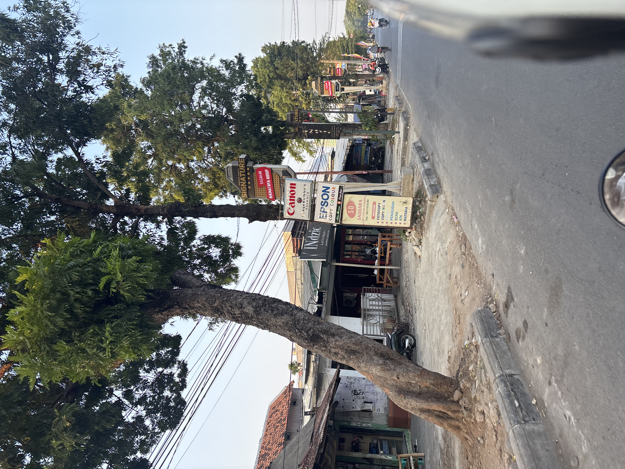 The image depicts a street scene in Cirebon, Indonesia, likely in a commercial area. The foreground shows a paved road with a concrete curb and sidewalk. A large tree with a thick, gnarled trunk leans into the scene from the left. On the right side of the road, various shops and businesses are visible, indicated by signage. One prominent sign displays "SIGNATURE" in large letters, with smaller text below reading "RASAKAN NIKMATNYA HIDUP". Another sign features the Canon logo and "IMAGERUNNER ADVANCE", while below it, an EPSON sign reads "COPY COLOUR". Further down the street, more businesses line the road, with some people visible inside or near the establishments. Several motorbikes are parked along the sidewalk, and a few are in motion on the road, suggesting active commerce and transportation. The architecture of the buildings appears to be a mix of older structures with tiled roofs and more modern shop fronts. The sky is clear and bright, indicating daytime. The overall impression is of a bustling urban environment.