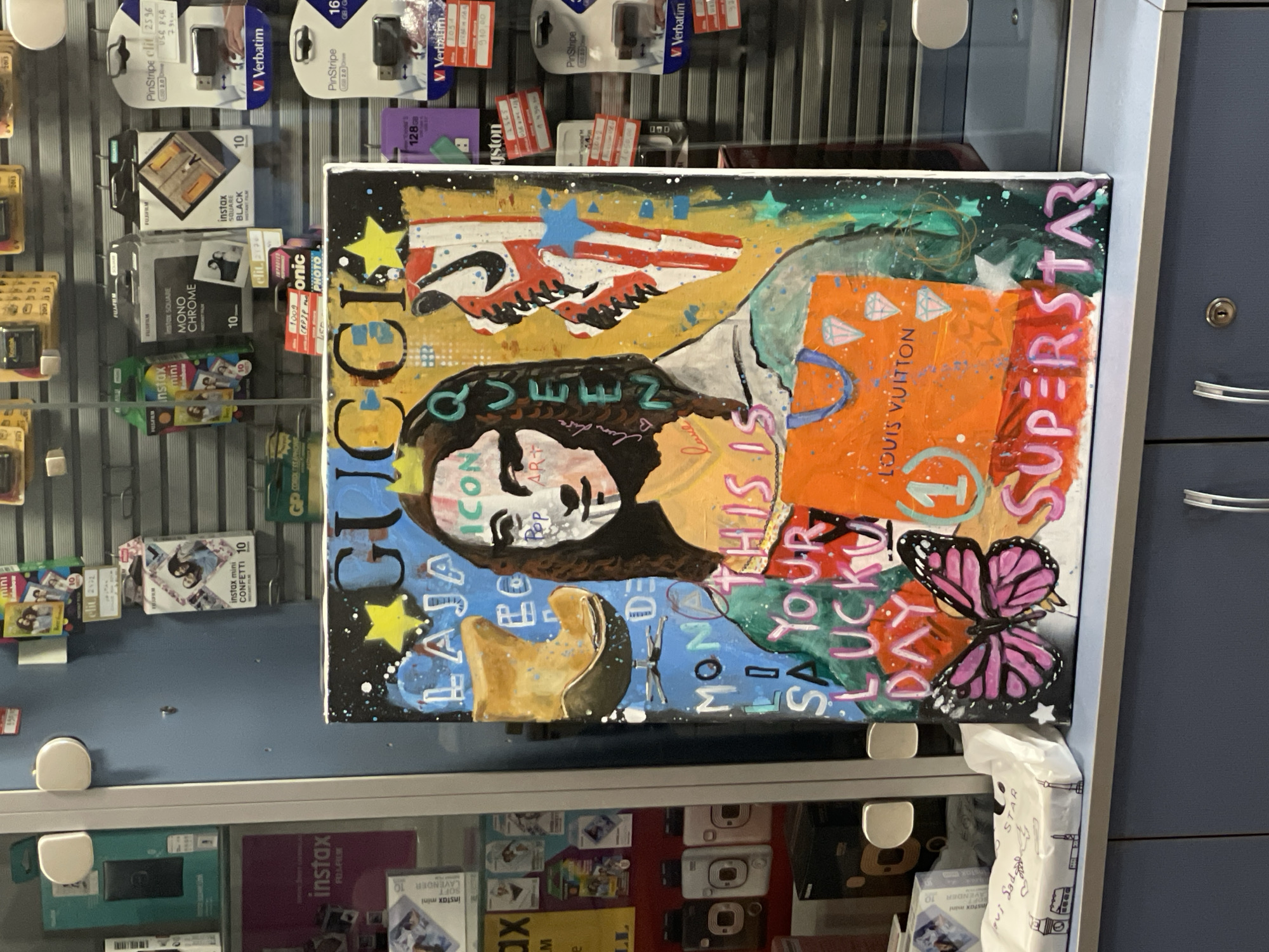 The image shows a painted canvas on display in what appears to be a retail setting. The canvas is positioned within a glass display case, partially obscuring shelves stocked with various products, including Verbatim USB drives, Fujifilm Instax film packs, and camera accessories. The painting features a stylized collage with images and text. Notable elements within the painting include the word "GUCCI" vertically stacked, a portrait-like image of a figure, a Nike swoosh with an American flag pattern, a Louis Vuitton shopping bag, a butterfly and various other words and decorative elements. A blue cabinet with chrome handles is visible adjacent to the display case. It is impossible to verify the location in Novi Sad, Serbia from the information available in the image.