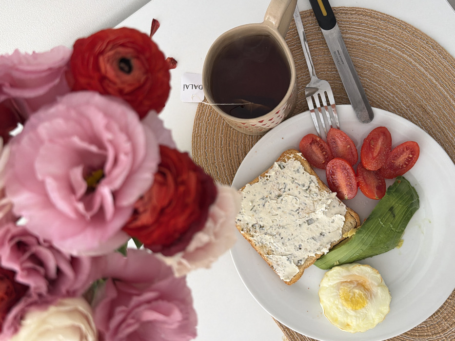 The scene depicts a breakfast setting on a white surface, presented as if located in Tel-Aviv, Israel. A white, round plate occupies the central lower portion, containing a single slice of toasted bread topped with a creamy, herb-infused spread, several slices of green avocado, approximately six halved red cherry tomatoes, and one fried egg with a visible yolk. To the right of the plate, a silver-colored four-tined fork and a silver-bladed knife with a black and yellow handle are laid on a woven, light brown placemat. The knife blade displays the inscription "ARCOS ALBACETE SPAIN." A beige or light brown ceramic mug containing a dark liquid, presumably tea, with a white tea bag tag labeled "DALAI" protruding, rests on the placemat to the right of the cutlery. The upper portion of the image is dominated by an out-of-focus bouquet of fresh flowers, including large pink blooms (likely Lisianthus) and deep red blooms (likely Ranunculus). The overall environment is illuminated, suggesting an indoor, daytime setting. No individuals are present in the image.
