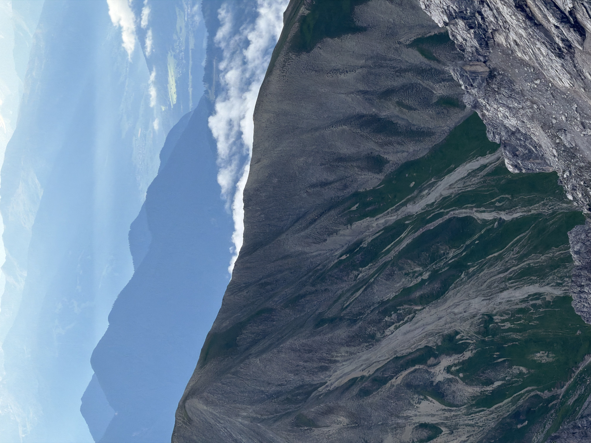 The image depicts a mountain landscape, likely from a high vantage point, looking out over successive mountain ranges. In the foreground, a steep, rocky slope with patches of green vegetation dominates the lower right quadrant. This slope transitions to a larger, darker mountain face that occupies the majority of the left and central portion of the image, characterized by steep scree slopes and sparse green growth.
In the middle ground, a valley or lower mountain range is visible, partially obscured by a layer of white, fluffy clouds. Beyond this, further mountain ranges fade into a hazy blue, indicating significant distance and atmospheric perspective. A hint of a settlement or scattered buildings can be discerned in the valley, suggesting a human presence within the expansive natural scenery. The sky above is a pale blue, with some wisps of white clouds. The overall impression is one of vast, rugged, and distant mountain scenery.
While the image itself does not provide specific visual cues to definitively place it in Garmisch-Partenkirchen, Germany, such a landscape is consistent with the Alpine regions surrounding the town, known for its dramatic mountain vistas.