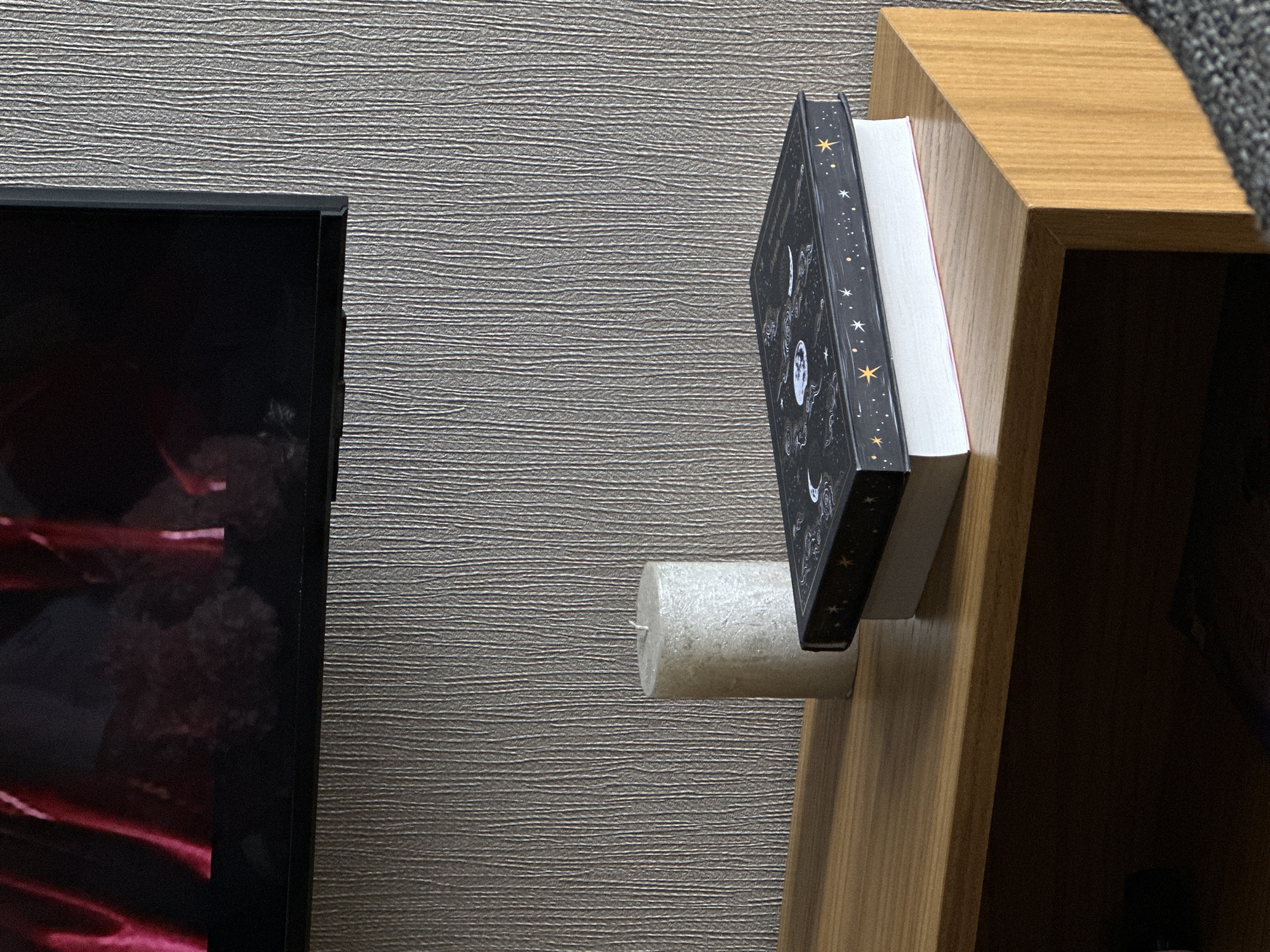 The image shows a close-up of a wall with a television on the left and a wooden shelf on the right. The television screen is dark, with blurred red and black shapes visible. The wall has a textured, gray-brown surface. On the shelf, a cylindrical white candle supports two books. The books are stacked vertically; the top book has a black cover decorated with white and gold celestial patterns (stars, moons). The second book has a plain white cover. The wooden shelf is light brown with a distinct wood grain. The background of the shelf interior is dark, indicating a shelf compartment. The image contains no people or actions. The location is not discernible from the available information.