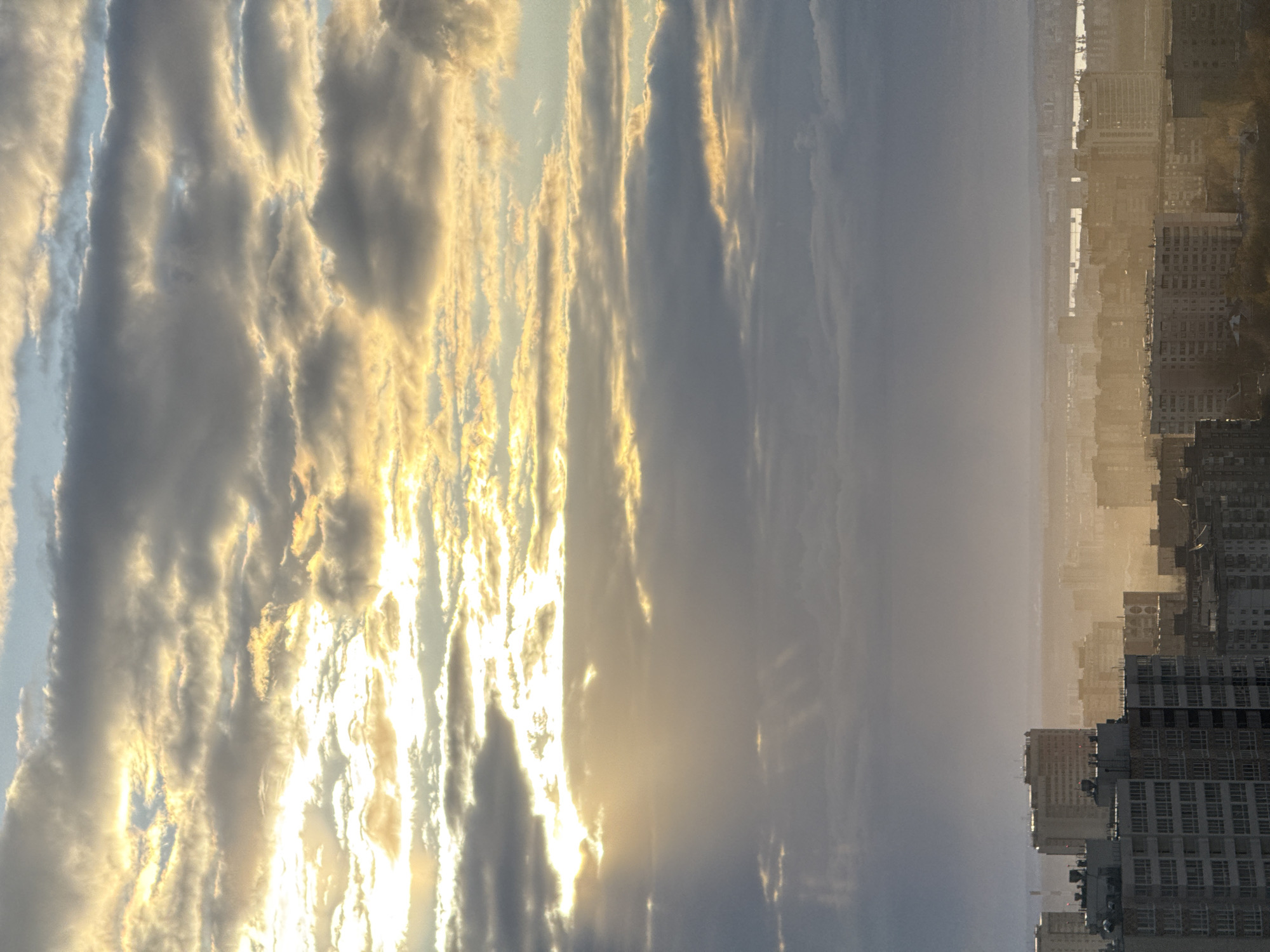 The image depicts a hazy cityscape in Perm, Russia, viewed from an elevated vantage point, likely a high-rise building. The scene is bisected, with the left portion showing a dramatic, cloud-filled sky illuminated by the setting sun. The clouds are various shades of gray and white, with golden edges where the sunlight catches them. Rays of light penetrate through the clouds, adding to the dramatic effect. The right portion of the image features the city of Perm, shrouded in a haze. Buildings of varying heights are visible, with taller apartment blocks near the bottom of the frame (as oriented) and lower buildings further back, receding into the horizon. The city has a uniform, subdued color palette due to the haze. There are no people visible in the image, and no specific actions are occurring within the frame. The environment is predominantly urban with atmospheric elements creating a sense of distance and visual obstruction.