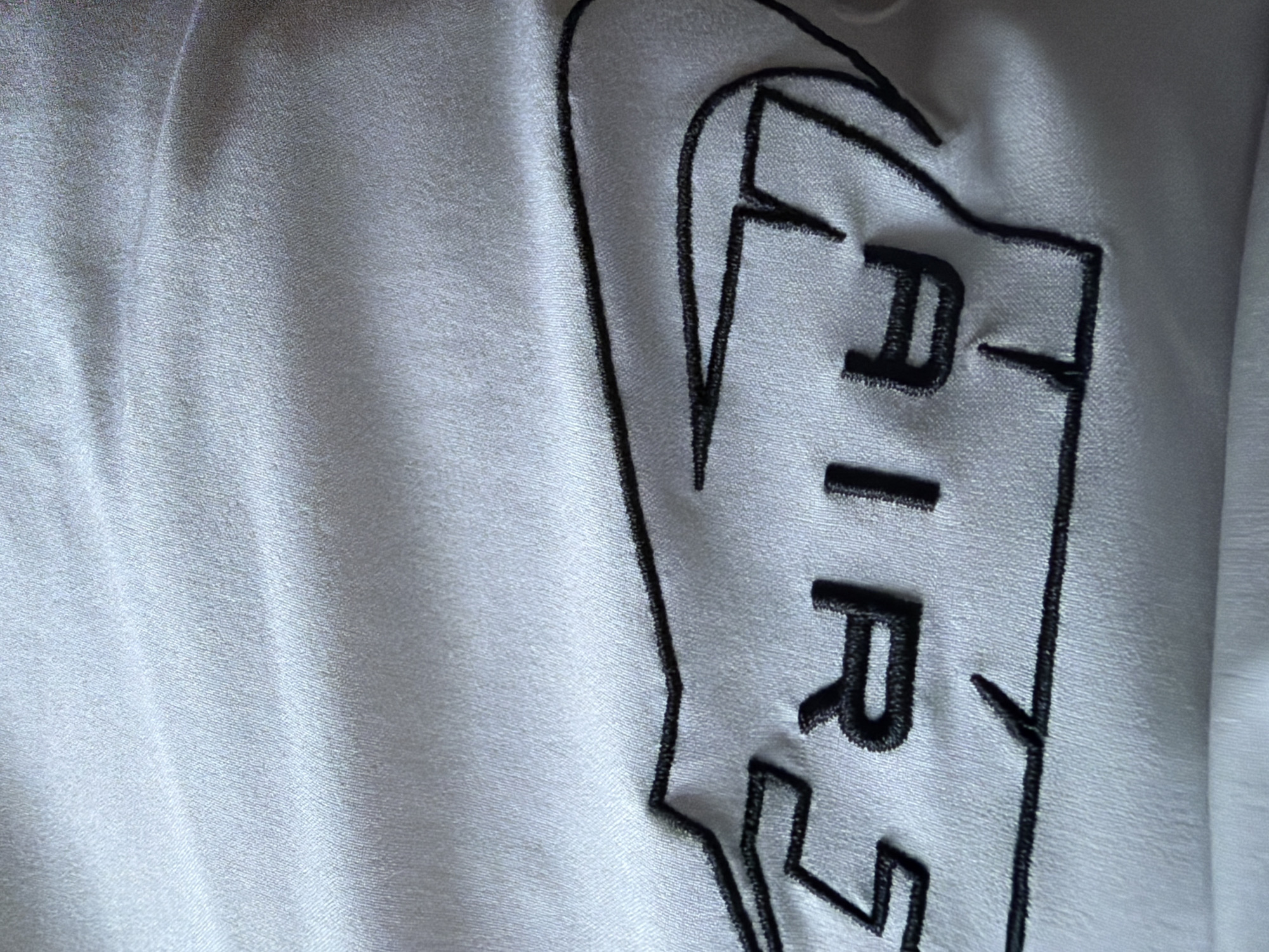 A close-up, high-angle shot captures a detail of a light-colored, possibly white or silver-grey, fabric. The material exhibits a subtle sheen and soft folds, characteristic of satin or a similar smooth textile. Dominating the right side of the frame is an embroidered design rendered in black thread. The prominent visible text spells "AIR," oriented vertically with each letter (A, I, R) clearly defined in a bold, block-style font. This word is encased within a thick, irregular black outline that suggests a larger, stylized logo or shape, potentially resembling a wing or a distinct brand identifier. The intricate texture of the embroidery, showing tightly packed stitches, is visible. Soft, diffused lighting highlights the fabric's sheen and the raised quality of the embroidery, creating gentle shadows along the folds. The image is a static portrayal of a fabric detail, likely from an item of clothing or an accessory, located in Razlog, Bulgaria.
