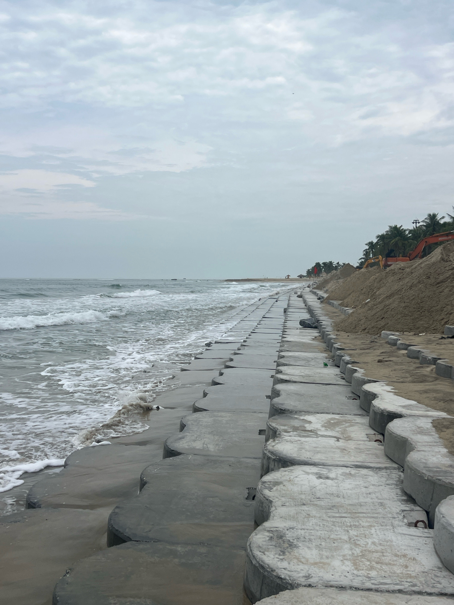 A view of the coast of Hoi An, Vietnam, shows a line of concrete wave-breaking blocks along the beach. The blocks are interlocking and have a curved shape, forming a barrier against the sea. The ocean is calm with small waves lapping at the shore. Beyond the blocks, a sandy embankment has been created, with piles of sand and construction equipment visible. Several palm trees dot the landscape in the background, suggesting a tropical or subtropical climate. The sky is overcast with clouds. The scene suggests ongoing coastal protection or construction work.
